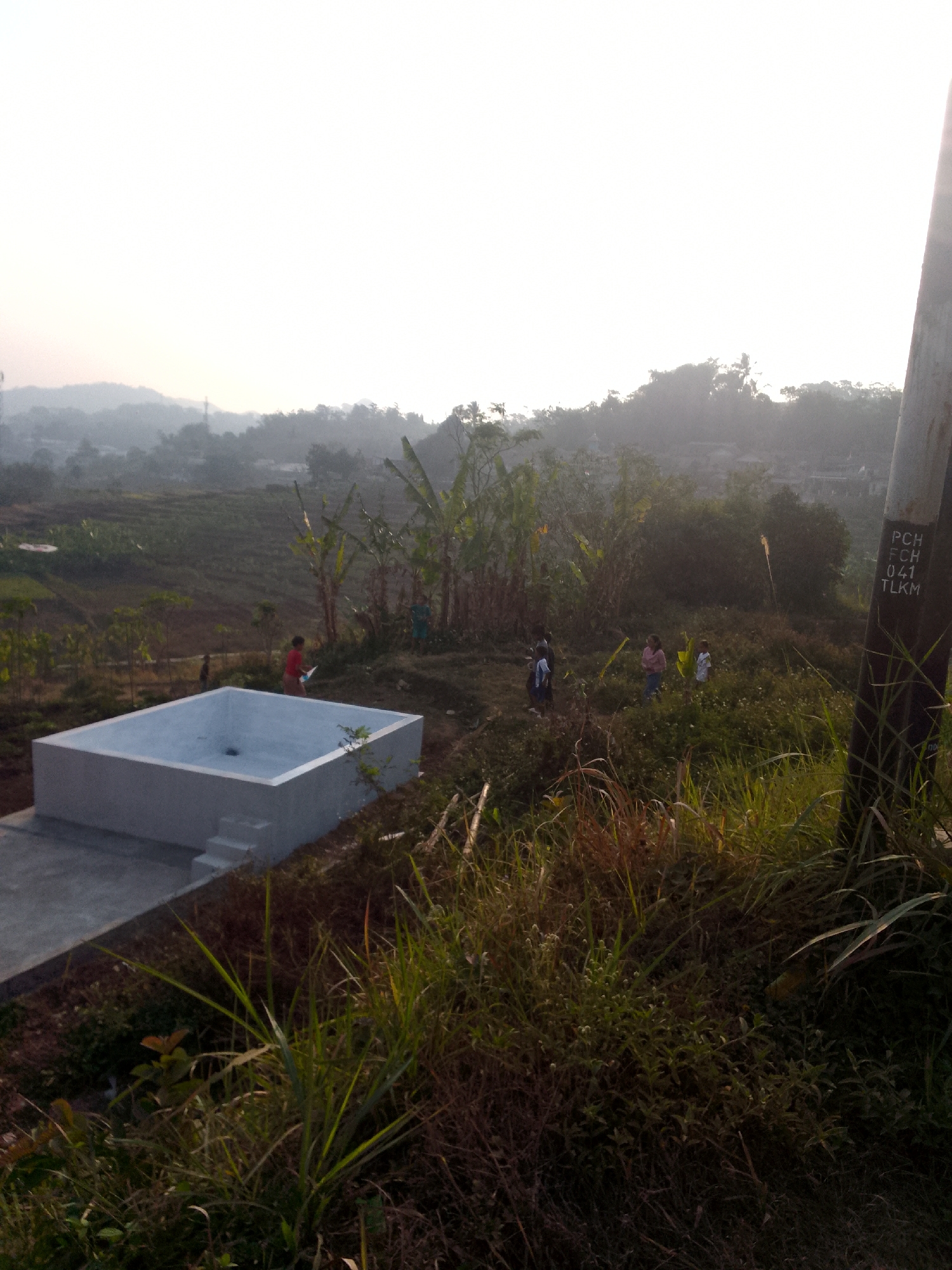In a rural setting, possibly in Padalarang, Indonesia, a concrete rectangular water tank with steps is situated on an elevated area. Several people, including children, are scattered throughout the scene, some walking and others standing. The environment is characterized by lush vegetation, including tall grasses and trees, with a hazy, mountainous landscape visible in the background. A wooden utility pole with markings is present on the right side of the image. The overall scene suggests a daytime activity in a developing or undeveloped area.
