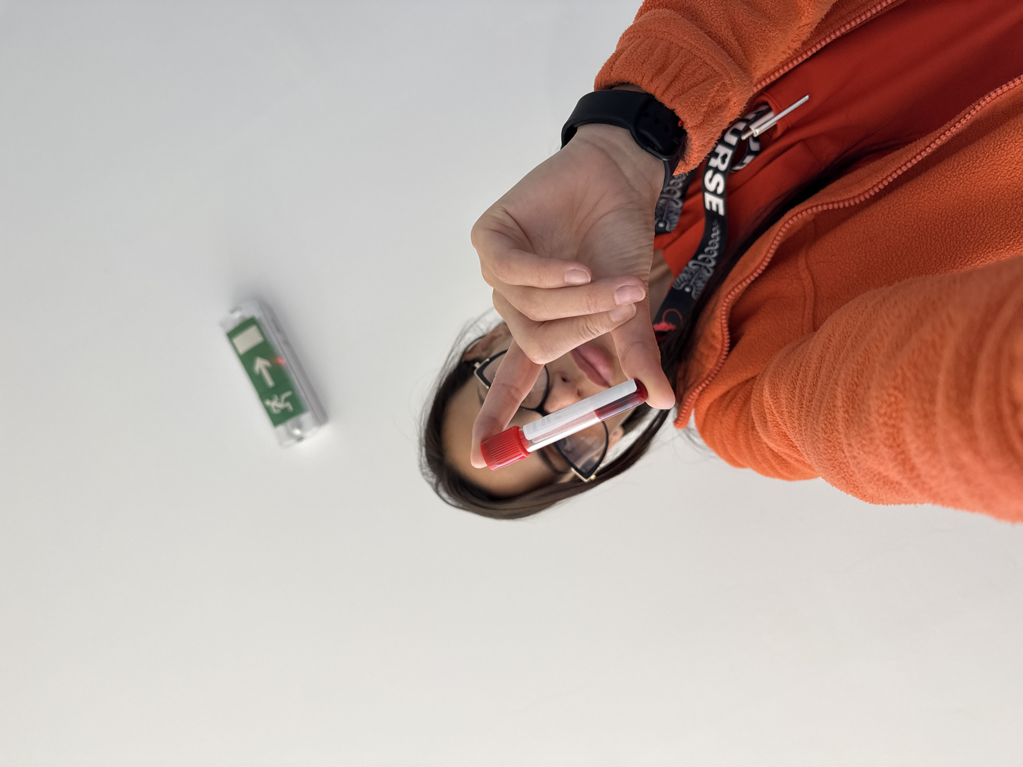 A person wearing glasses and an orange fleece jacket is holding a blood sample vial in their hand. The vial has a red cap and contains a red fluid, likely blood. A black smartwatch is visible on the person's wrist. To the left of the person, an emergency exit sign is on a white surface. The location is unknown.