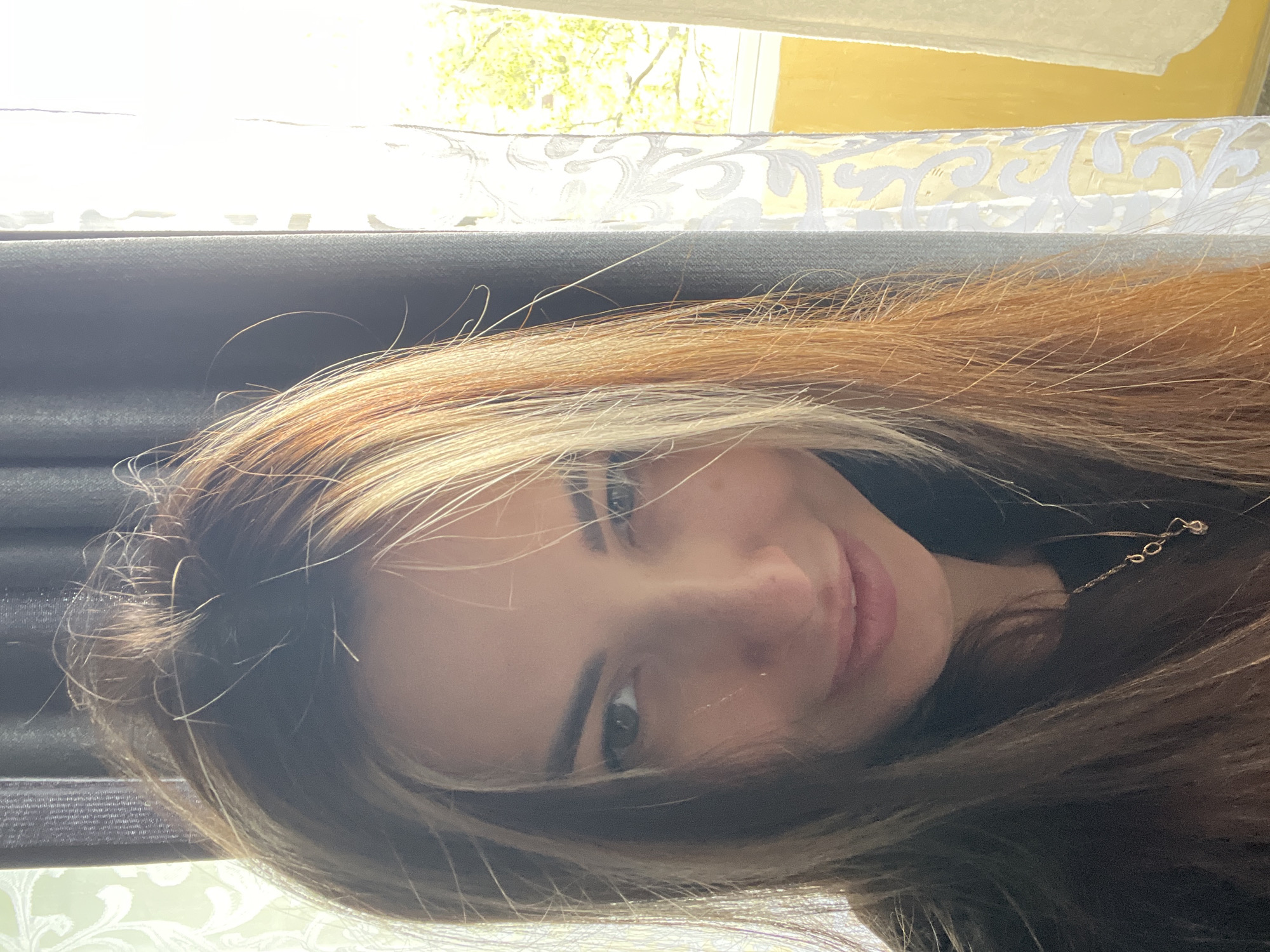 A woman with long, brown hair is positioned in front of a window. The window features a sheer white curtain with a decorative pattern and a yellow wall is visible outside. The city is Unknown, Unknown.
