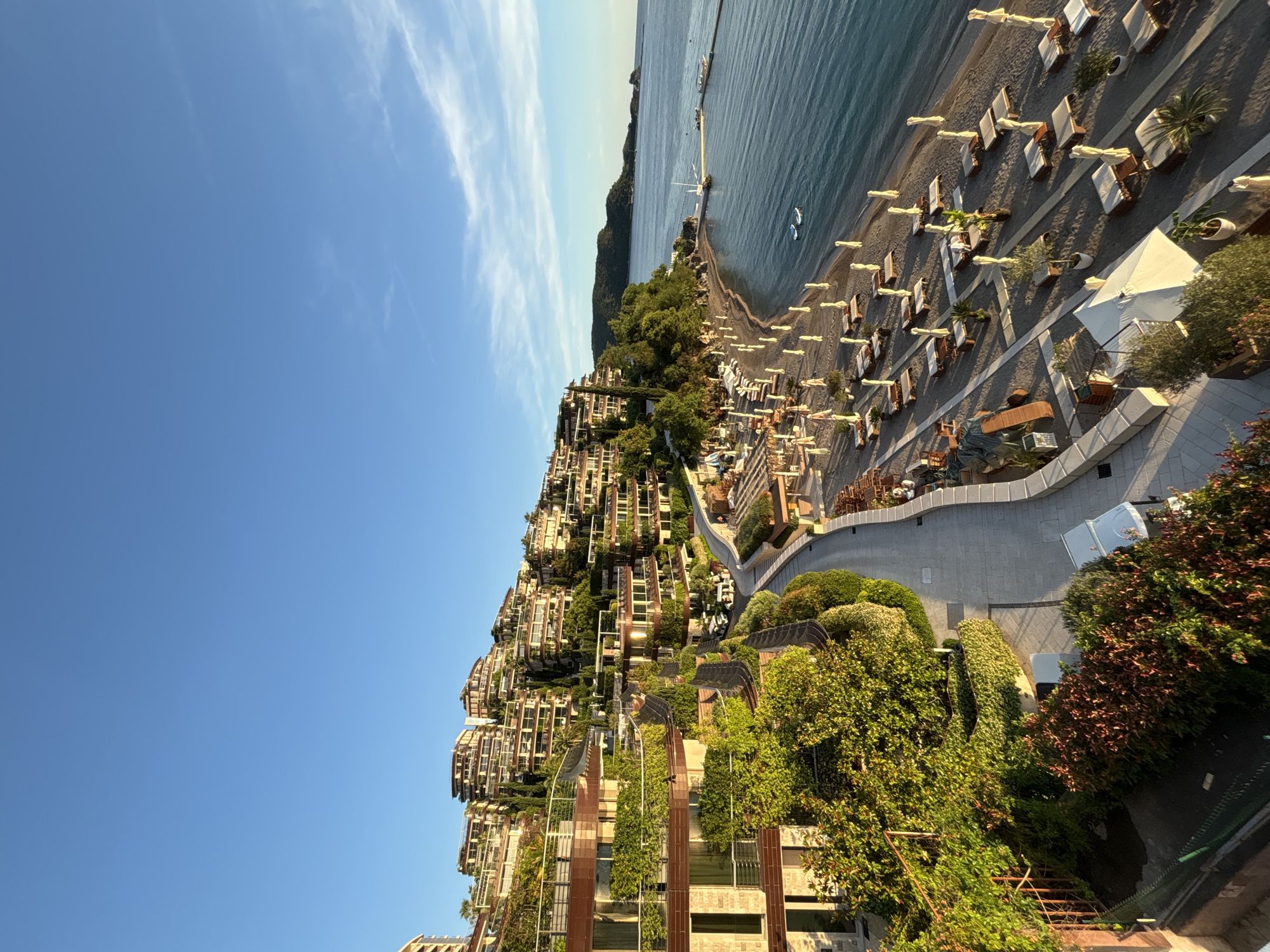 The image presents a serene, panoramic view of a high-end coastal resort or residential area in Budva, Montenegro. The scene is dominated by a clear blue sky with a few wispy white clouds at the top. Below, a calm, blue-green sea meets a long stretch of dark-sanded or pebbled beach. A pier or jetty extends into the water at the far end of the beach, and two small white boats are visible further out in the sea.
The beach is meticulously arranged with numerous empty sun loungers and umbrellas, predominantly white or light-colored, suggesting an exclusive vacation spot. Small palm trees and potted plants are dotted along the beachside. A large white tent-like structure stands on the beach, likely serving as an amenity for guests.
Behind the beach, the hillside is densely covered with modern multi-story buildings. These structures are designed with extensive "green architecture," featuring numerous balconies and terraces adorned with lush green vegetation, including climbing plants, shrubs, and small trees, creating a harmonious blend with the natural environment. The buildings exhibit a mix of dark brown and lighter-colored facade panels. A winding, paved road or pathway gracefully snakes through these verdant buildings, connecting them to the beachfront. Manicured bushes and dense foliage further enhance the green landscape.
No people are visible, lending a quiet, undisturbed ambiance to the scene. The bright, clear sunlight and visible shadows suggest it is either late morning or early afternoon under perfect weather conditions. The overall impression is one of luxurious tranquility and natural beauty, indicative of a premium coastal destination. There is no visible text in the image.