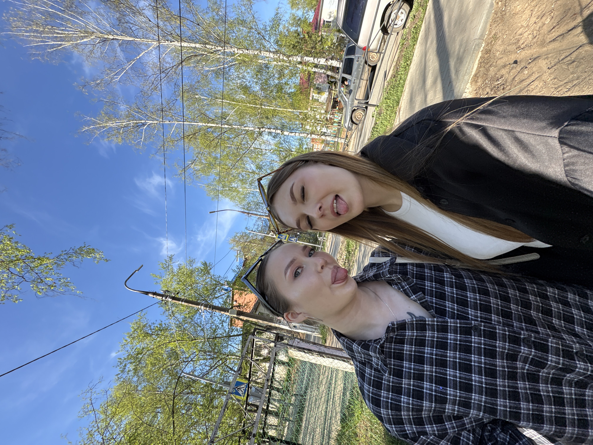 Two young women pose for a selfie on a sunny day. The location appears to be a street in the city of Chaikovsky, Russia. The woman in the foreground wears a black and white checkered shirt, with sunglasses resting on her head. A silver necklace is visible, and a tattoo is partially visible on her upper chest. She is sticking out her tongue. The woman behind her wears a black blazer over a white top and sunglasses on her head. She is also sticking out her tongue. In the background, trees with green foliage, a sidewalk, and a parked silver van can be seen. A utility pole and wires are also visible. A fenced area and a building are also in the background. The sky is blue with some clouds.