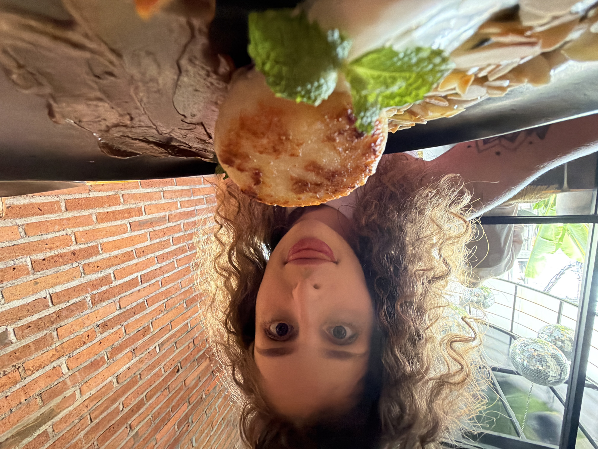 A woman with curly brown hair is leaning towards the camera, sticking her tongue out. She is in what appears to be a restaurant with a brick wall in the background. In front of her is a black plate with food on it. The food includes a round, golden-brown pastry or cake, thinly sliced almonds, a sprig of mint, and a dark, textured sauce. The overall lighting suggests it might be daytime. The presence of disco balls visible through a window hints at a potentially lively or entertainment-focused establishment. There are no other people clearly visible in the frame. The scene is shot from a low angle, making the food appear prominent. The location is specified as North Kuta, Indonesia.