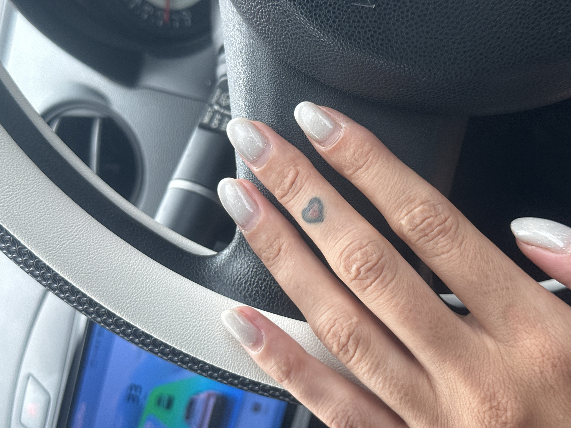 A person's hands are resting on a car's steering wheel. The nails are long, oval-shaped, and painted with a pale, glittery polish. A small heart tattoo is visible on the ring finger. The steering wheel is black leather with a textured gray and white trim. In the background, a car's dashboard display shows a navigation screen with the number "33". The scene appears to be inside a vehicle, likely parked or in traffic. There is no information to place this image in Ko Samui District, Thailand.