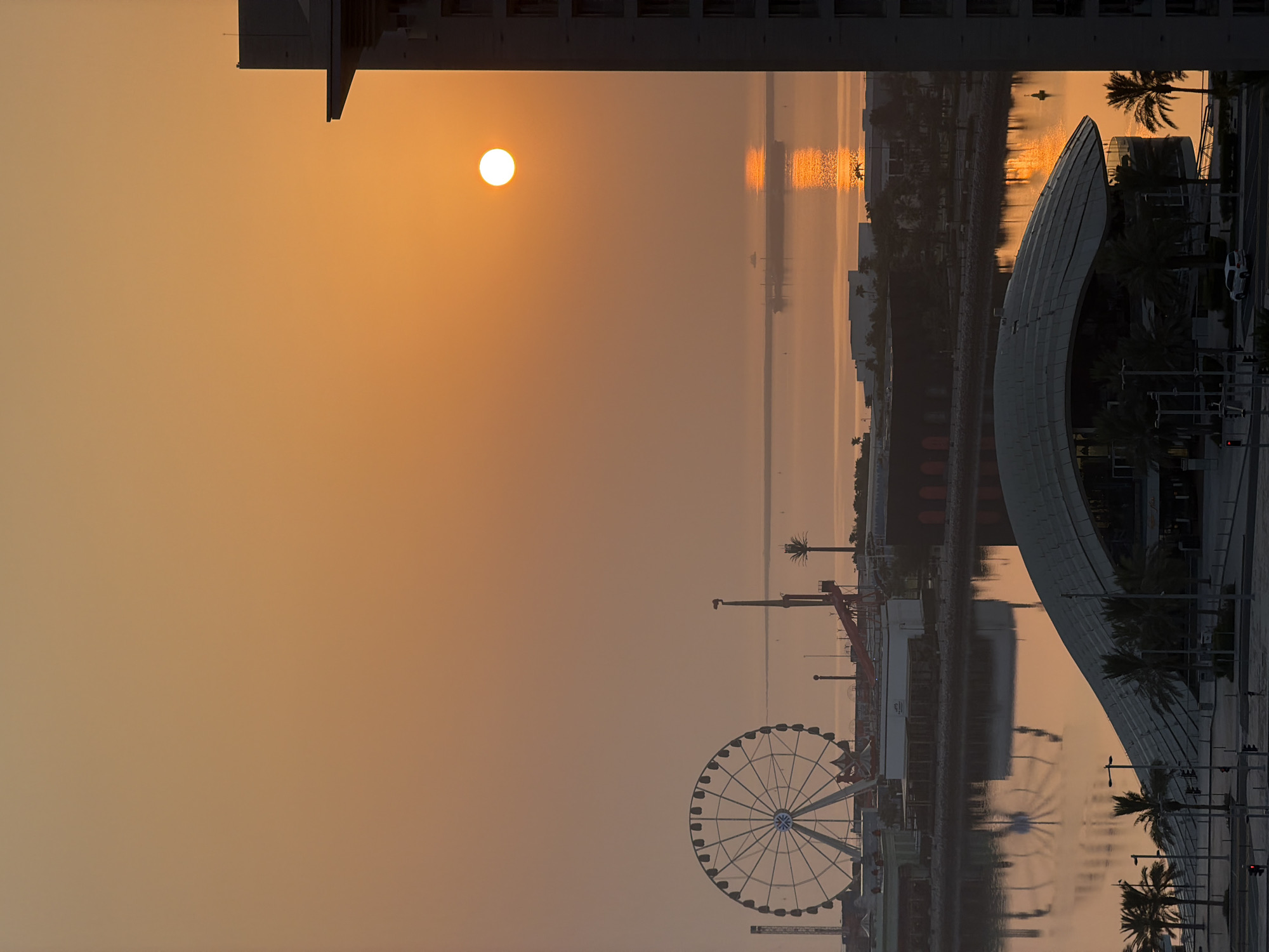 A wide shot captures a tranquil scene at sunrise in Lusail, Qatar. The sky is a soft orange hue, gradually transitioning to a lighter shade towards the horizon. A bright, round sun hangs in the upper right quadrant, casting a warm glow and a shimmering reflection on the calm water. In the foreground, the sleek, curved roof of a modern building is visible, surrounded by palm trees. Across the water, a Ferris wheel stands prominently, hinting at an amusement park. Buildings and infrastructure line the waterfront, with a boat visible in the distance. The image conveys a sense of peace and urban development against a natural backdrop.