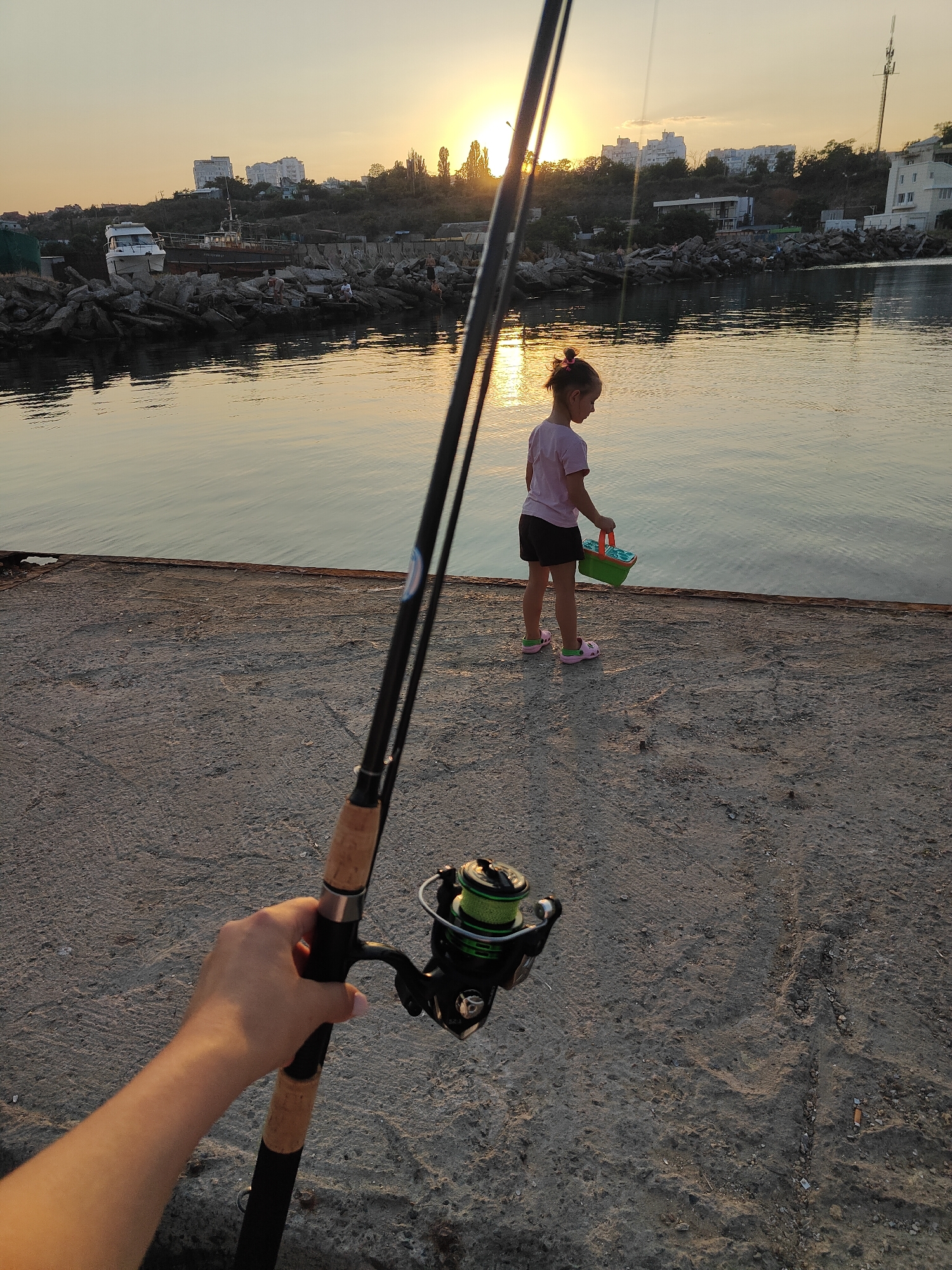 A young girl stands on a concrete pier by the sea in Chornomorsk, Ukraine, holding a green bucket. She is wearing a pink t-shirt and black shorts. Behind her, the sun is setting over the water, casting a warm glow on the scene. In the foreground, a fishing rod is held by a person whose hand and arm are visible. In the background, several buildings are visible on a hill overlooking the sea, and there are some boats docked nearby. The overall atmosphere is peaceful and serene, with a hint of anticipation for fishing.