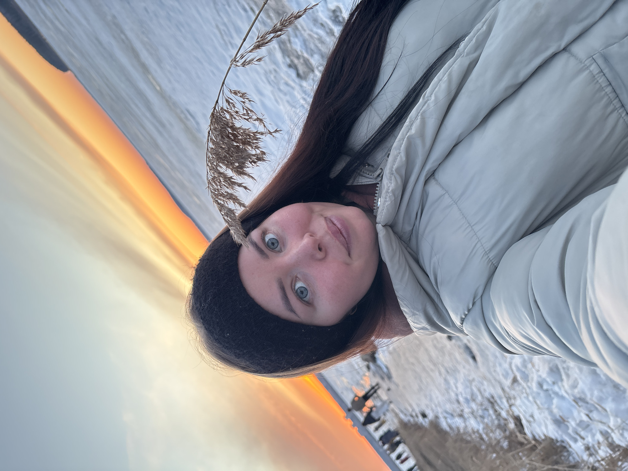 The image is a selfie of a woman wearing a black knit hat and a light grey puffer jacket. Her hair is long and dark brown. She has light skin and blue eyes. A stalk of dry grass is near her face. In the background, there is a body of water, possibly a lake or river, with ice or snow on the surface. The sky has a gradient of colors ranging from orange near the horizon to light blue above. The setting could be near a city in Russia based on the request, but the visible details do not definitively confirm the location as Izhevsk, Russia. The woman appears to be outdoors, likely near the body of water, possibly during sunset. Some reeds are also visible in the lower right.