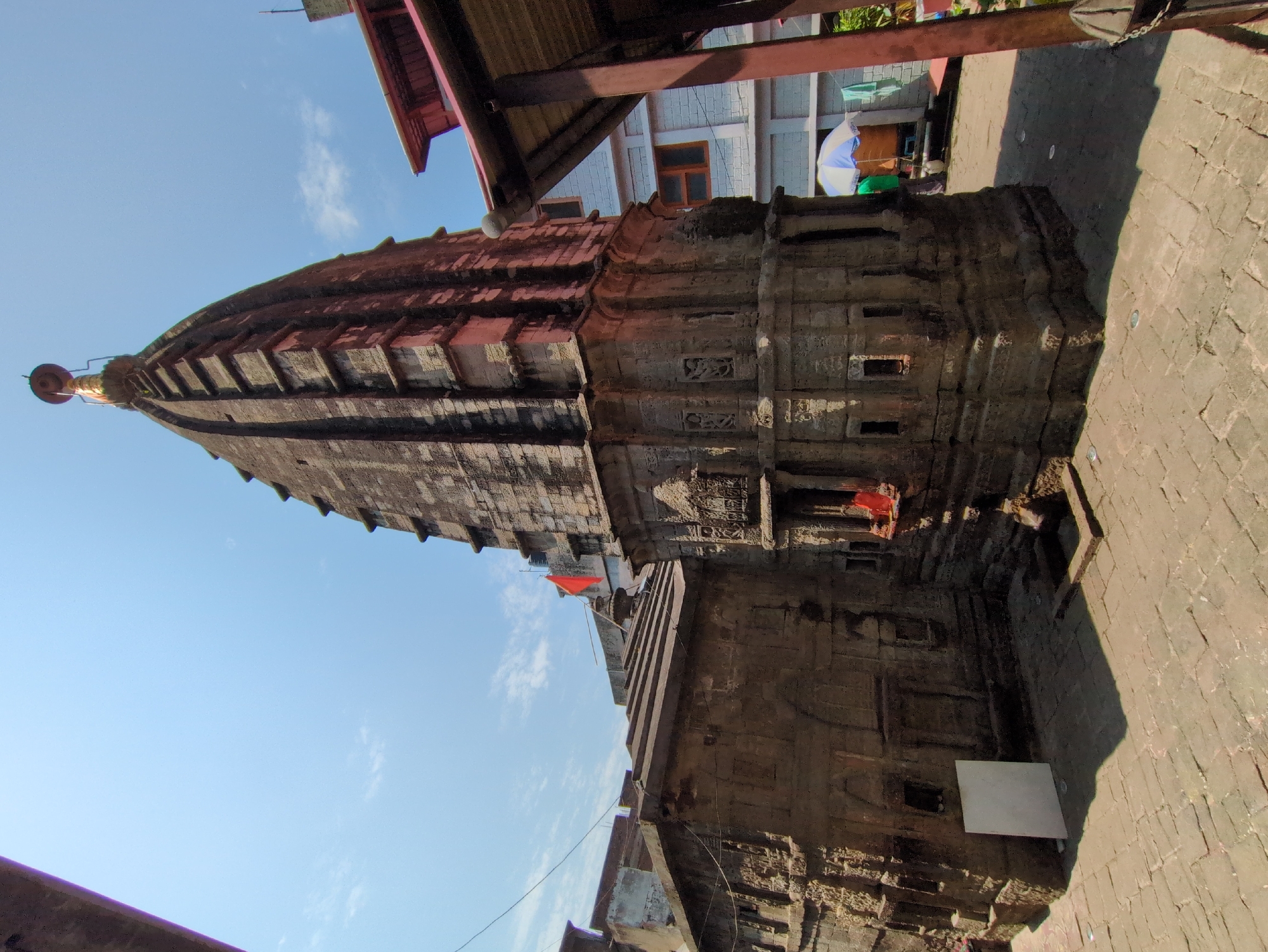 The image depicts a stone temple in Mandi, India, under a clear blue sky with a few wispy clouds. The temple, made of dark, weathered stone, has a tiered, pyramidal roof and intricate carvings on its facade. A red flag is visible near the top of the temple. In the foreground, the ground is paved with stone tiles, and shadows are cast by the structures. To the right of the temple, a modern building with red roofing is partially visible, along with a colorful umbrella and some draped cloth. The overall scene suggests a historic religious site within a populated area.