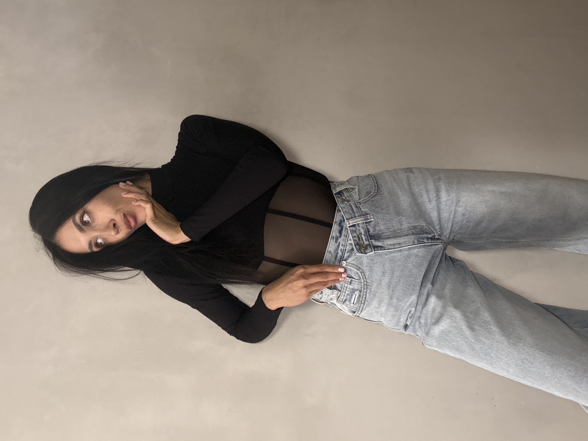 A woman with long, dark hair stands with her back to a neutral-colored wall. She is wearing a black, long-sleeved turtleneck top with sheer paneling and light-wash, high-waisted jeans. Her left hand is resting on her chin, and her right hand is tucked into the front pocket of her jeans. She is looking to her right, with her head tilted. The image does not contain any information that would indicate a location in Votkinsk, Russia.