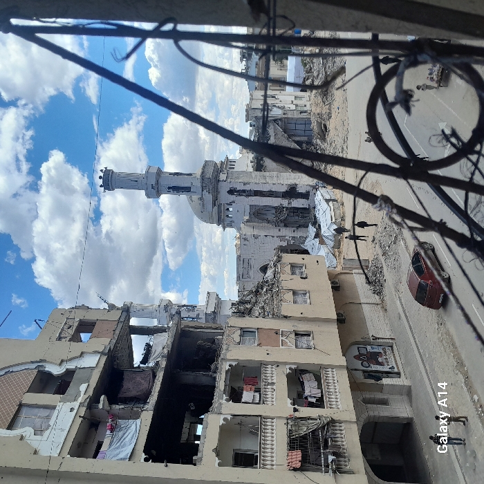 This image depicts a severely damaged urban landscape in Khan Yunis, Palestinian Territory, viewed from an elevated perspective through a metal grille.
In the foreground, an out-of-focus, dark-colored metal grille or fencing runs diagonally across the right side of the frame, partially obscuring the view.
To the left, a multi-story building, beige in color, exhibits extensive structural damage. Multiple floors are exposed with exterior walls collapsed or missing, revealing interior spaces filled with rubble. Balconies on several levels, some with decorative white balustrades, show clothes hanging, including a white sheet and other garments, indicating partial habitation or presence of personal belongings. The ground floor of this building features a partially visible sign that includes "MESSA GAMES" in English.
In the mid-ground, a prominent mosque, light-colored and featuring a tall minaret with a crenellated top, stands heavily damaged. The main structure of the mosque behind the minaret has significant sections of its walls missing, exposing interior spaces. Rubble surrounds the base of the mosque and extends into the area between it and the damaged building on the left. To the left of the main minaret, a shorter, more extensively damaged minaret or tower is visible. Scattered white tarpaulins, possibly forming makeshift shelters, are present amidst the debris in the open area.
A paved road with a white center line runs across the lower mid-ground. A red sedan is parked or stationary on the left side of this road, facing away from the viewer.
Several individuals are present in the scene: two figures, appearing as men or older boys, stand near the curb on the left side of the road, close to the "Galaxy A14" watermark. Further into the scene, three individuals are walking across the debris-strewn ground in front of the mosque, heading towards the background. On the far right of the road, another individual stands next to a small, low cart or stand.
The sky is bright blue with scattered large white cumulus clouds, suggesting daytime conditions.