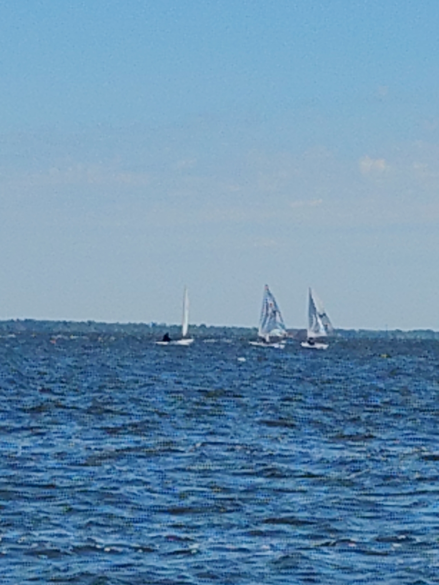 The image captures a serene daytime scene on a body of water, likely a river or lake, under a clear blue sky. Several sailboats are visible, navigating the choppy blue water. In the foreground, the water is a deep blue with whitecaps indicating moderate waves. Further out, a lone white sailboat with a single sail is sailing to the right, with a dark figure visible in its hull. To the right of the lone sailboat, two other sailboats are sailing closer together. The one on the left has a triangular sail with some markings, and the one on the right also has a triangular sail. Both of these sailboats appear to be larger than the lone sailboat and are also sailing to the right. The horizon line is low, with a faint strip of land or trees visible in the distance. The overall impression is one of a pleasant day for sailing. The presence of sailboats on a large body of water suggests a location that is a popular spot for recreational activities. While the provided context states the location is Cherkasy, Ukraine, specific landmarks or city features are not discernible in this image.