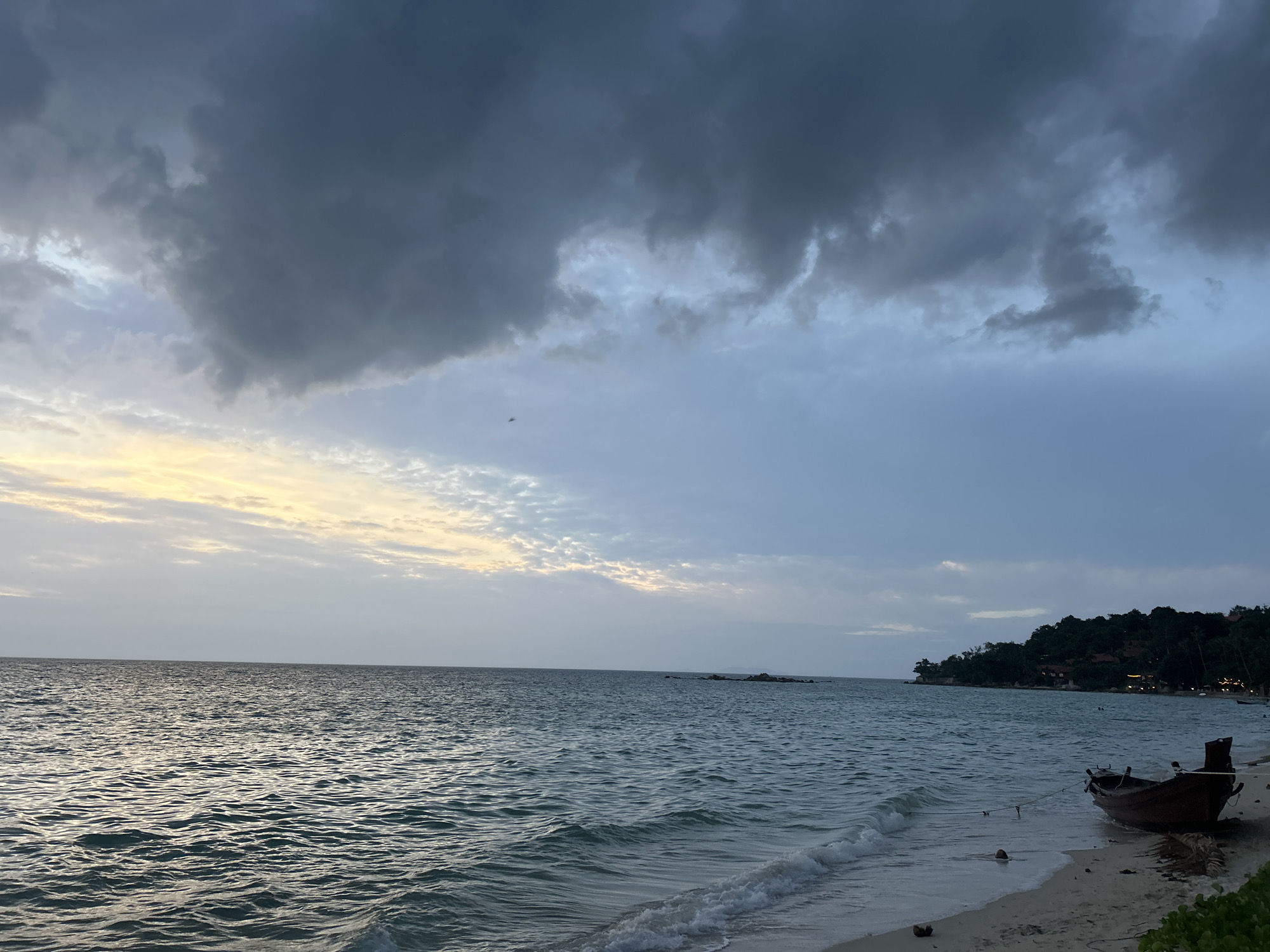 A twilight view of a sandy beach in Ko Pha-ngan District, Thailand, shows the ocean to the left and a tree-covered island in the distance to the right. Dark, heavy clouds cover the upper portion of the sky, with patches of lighter clouds and a sliver of yellow light near the horizon. The sea is a deep blue-green, with gentle waves lapping at the shore. A small, wooden boat is beached on the sand in the lower right corner, tied to a rope. The sand is light brown, with a few scattered coconuts and dried palm fronds. A few small lights are visible on the island in the distance.