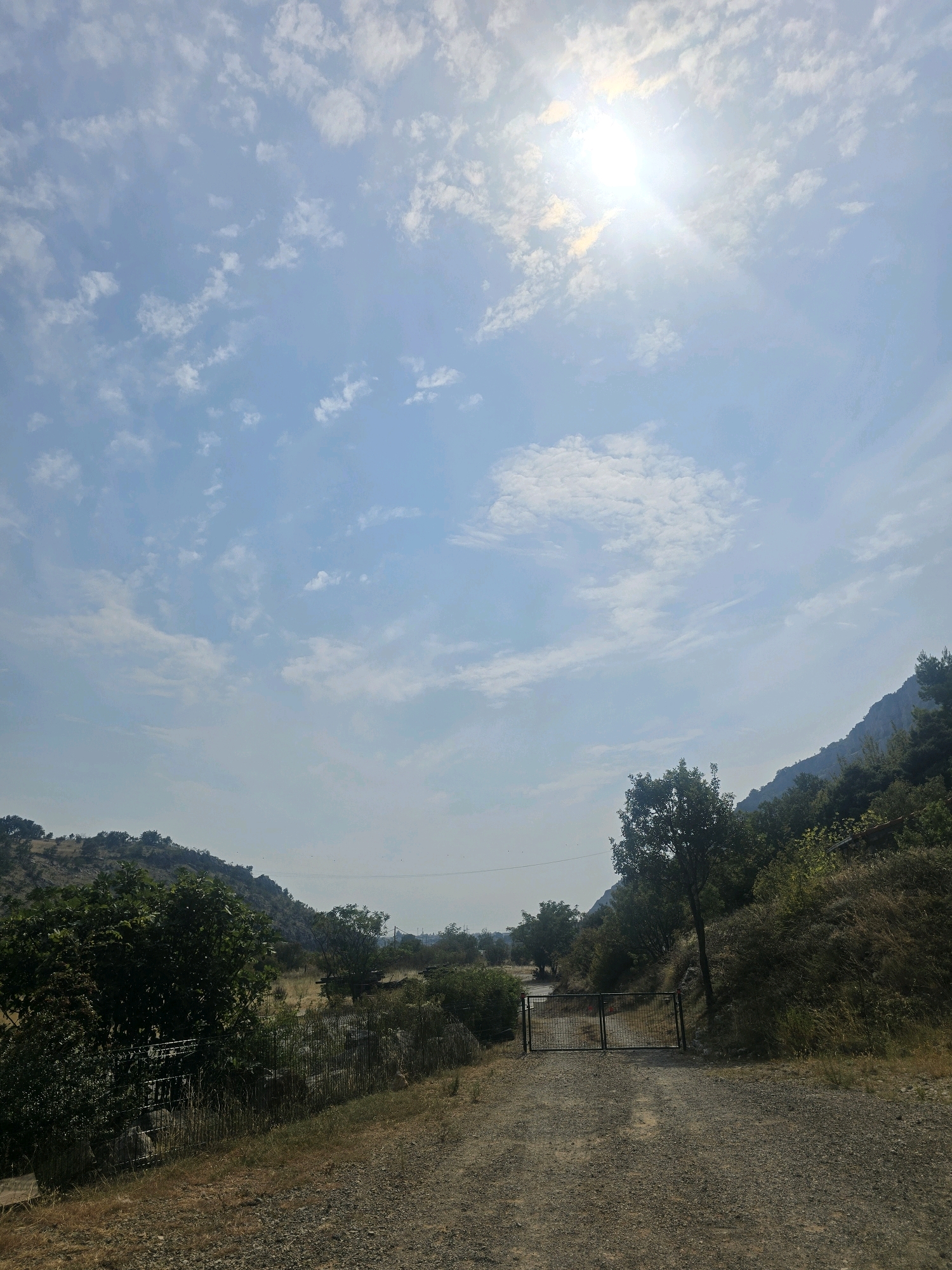 The image depicts an outdoor scene on a sunny day with a clear sky filled with scattered white clouds. The sun is visible and bright in the upper right quadrant, casting a strong light over the landscape. A dirt road or path leads towards a closed metal gate, flanked by trees and vegetation on either side. The path appears to be gravelly and uneven. To the left of the path, there is dense green foliage and a wire fence. The terrain on the right side of the path is a grassy slope covered with dry grass and scattered trees. In the background, hills or mountains are visible, covered with vegetation. The overall impression is of a rural or semi-rural environment, possibly a park, trail, or private property entrance. The image does not contain any people or specific landmarks that would definitively place it within the city of Podgorica, Montenegro.