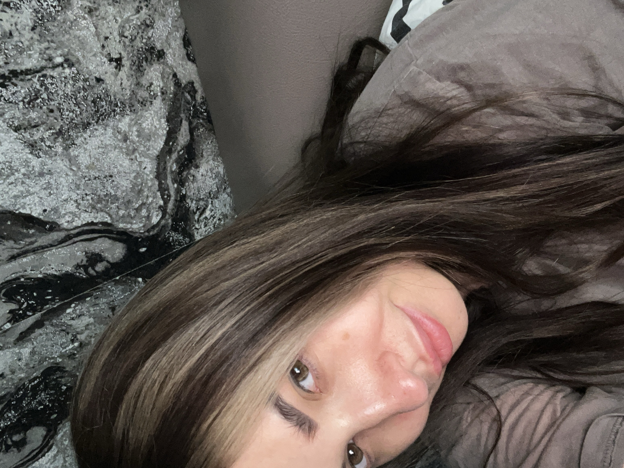 A close-up, overhead shot shows a person, likely female, reclining indoors. The subject's face is visible from the eyes to the mouth, tilted slightly upwards and to the right, framed by dark brown hair with lighter, possibly highlighted, streaks. She has brown eyes and her lips are slightly parted, forming a subtle, gentle smile or smirk. Her gaze is directed slightly away from the camera.
She appears to be resting on or beside two distinct fabric surfaces. To her left, a patterned material exhibits an abstract, marbled design in shades of grey, black, and white, with shimmering or textured elements. Behind her head and to her right, a plain, soft grey fabric, possibly a pillow or blanket, is visible. Her long, dark hair is spread across both textures. The setting suggests a casual, relaxed atmosphere, likely in a home environment like a bedroom or living room. There is no visible text in the image.