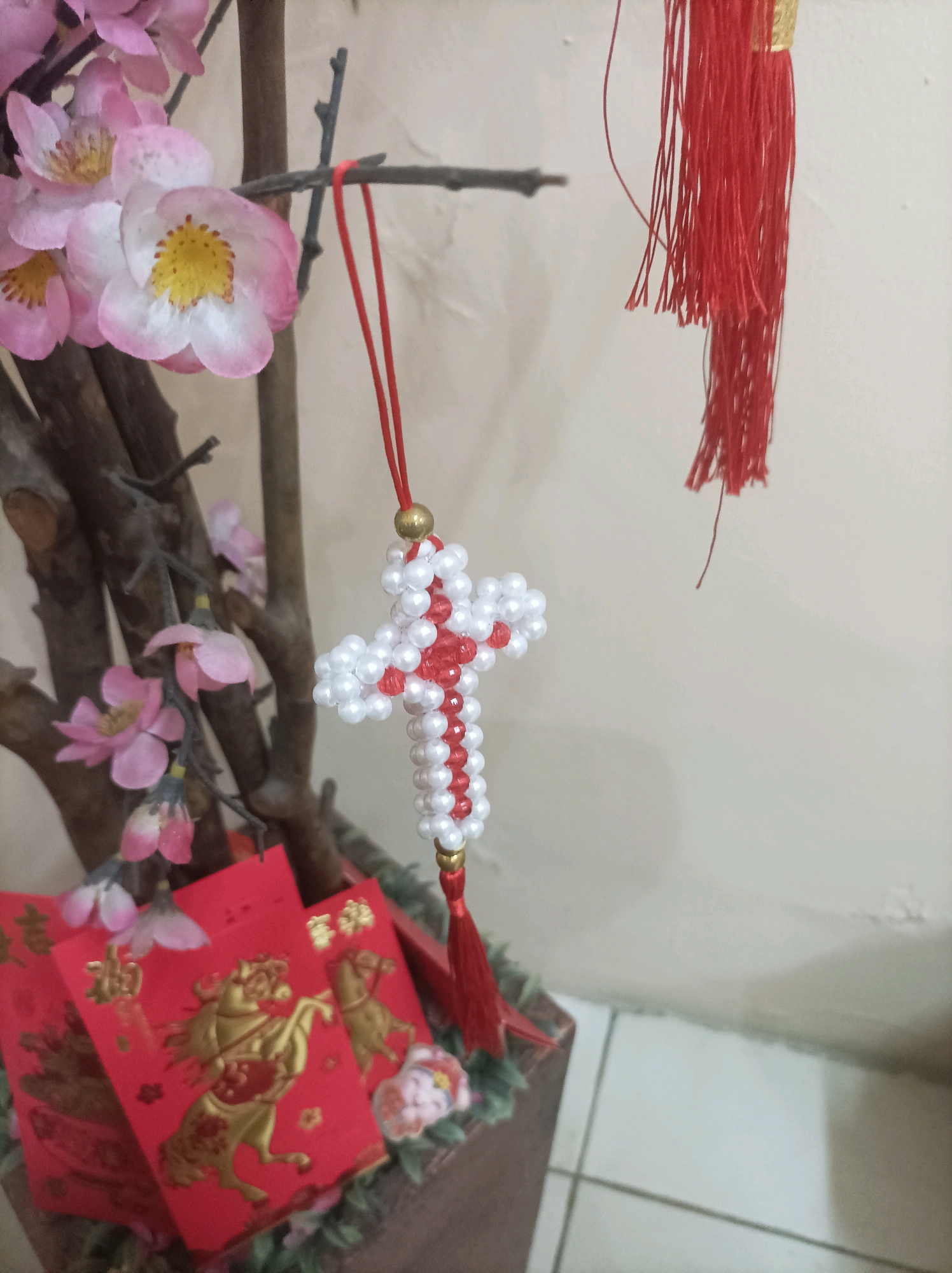 The image depicts a close-up of a decorative arrangement, likely for a festive occasion. In the foreground, several red envelopes with golden horse imagery are visible, suggesting a Chinese New Year or similar celebration. These envelopes are nestled amongst artificial pink flowers and branches, which are part of a larger decorative plant. Hanging from one of the branches is a beaded ornament shaped like a cross, alternating between white and red beads, with a red tassel at its base. A separate red tassel hangs from a branch above and to the right of the cross ornament. The background is a plain, light-colored wall. The lighting appears to be artificial, casting subtle shadows. The scene is indoors, likely in a residential setting. The specific location of Bekasi, Indonesia is not discernible from the visual information provided.