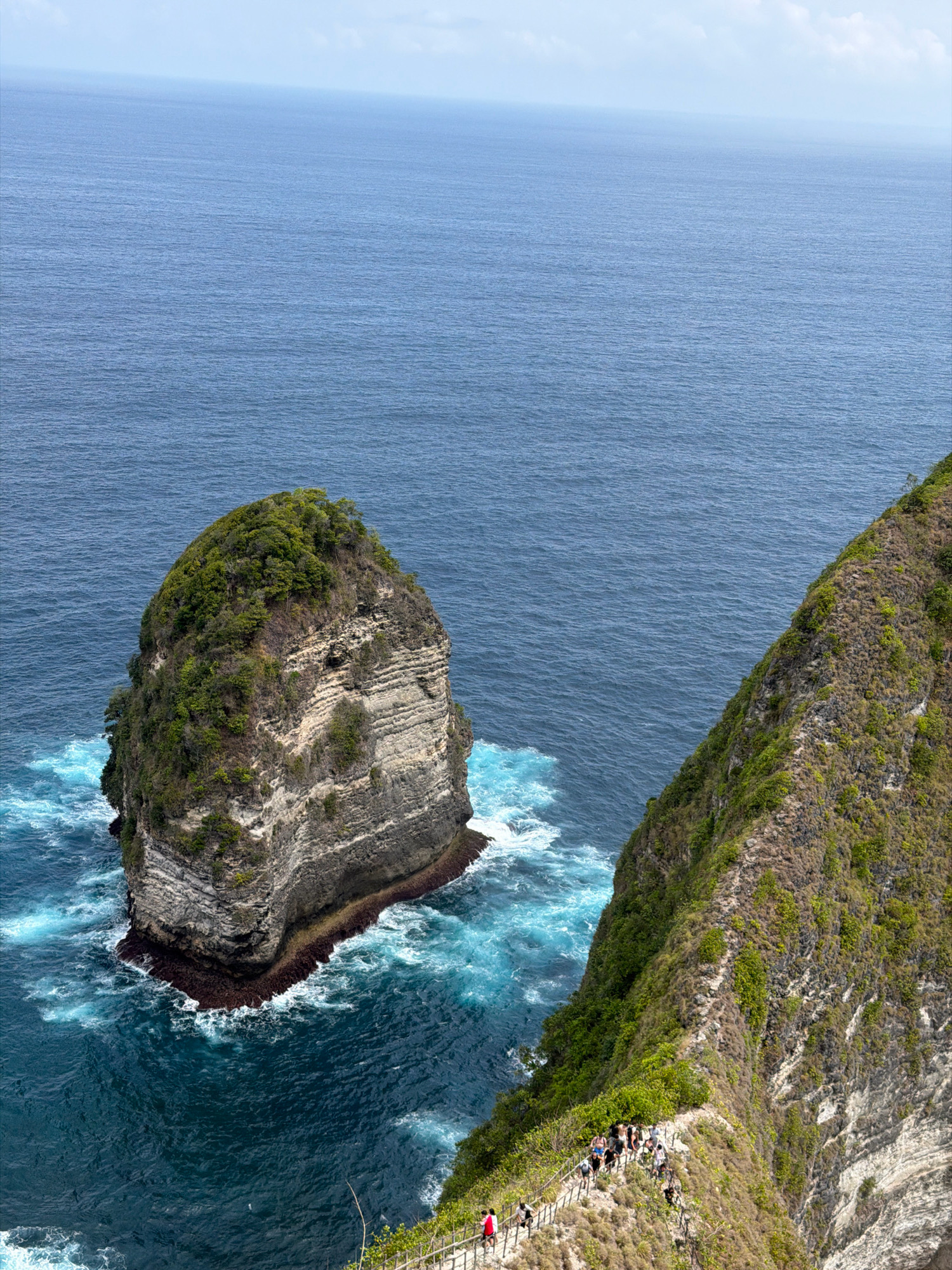 This is an aerial, high-angle shot of the Kelingking Beach cliff in Nusa Penida, Indonesia. The ocean is a deep blue with lighter turquoise waves crashing against the shore. On the left, a large, rocky island covered in lush green vegetation rises from the sea. To the right, the sheer cliff face slopes down, with a narrow path winding along its edge. A group of people, wearing casual clothing, are visible on the path, some walking, some standing. They appear to be tourists taking in the scenic view. The sky above is a pale blue with hints of clouds.