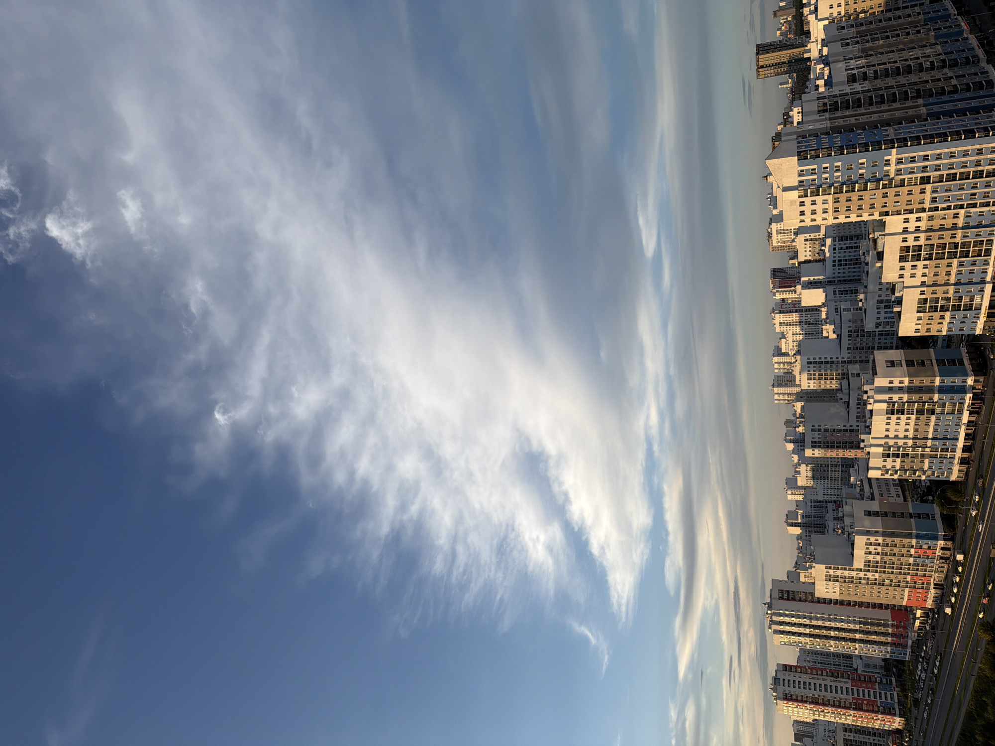 This is a high-angle, vertical shot of a residential cityscape at dusk, likely in Yekaterinburg, Russia. The scene is dominated by numerous tall, modern apartment buildings, characterized by their multicolored facades of white, blue, red, and gray. These buildings are densely packed together, forming a sprawling urban landscape. A road with cars is visible in the foreground, suggesting active traffic. Above the city, the sky is a gradient of deep blue at the top transitioning to lighter shades towards the horizon, filled with wispy, elongated clouds that are illuminated by the setting sun, casting a warm glow. The overall impression is one of a modern, developing urban area under a dramatic sky.