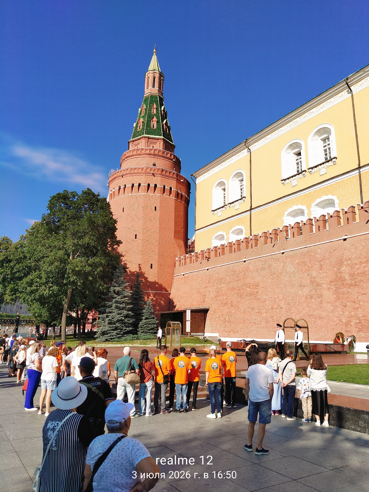 The image depicts a daytime scene in an urban environment, likely a historical site, featuring a prominent red brick tower with a green spire. In front of the tower, a group of people are gathered, some wearing matching orange t-shirts with white graphics. To the right of the tower, a yellow building with arched windows is visible, and a long red brick wall extends from the tower. Two individuals in white uniforms and hats stand guard near a memorial flame. The ground is paved, and trees are present in the background. The sky is clear and blue. A watermark with text is visible in the lower right corner.