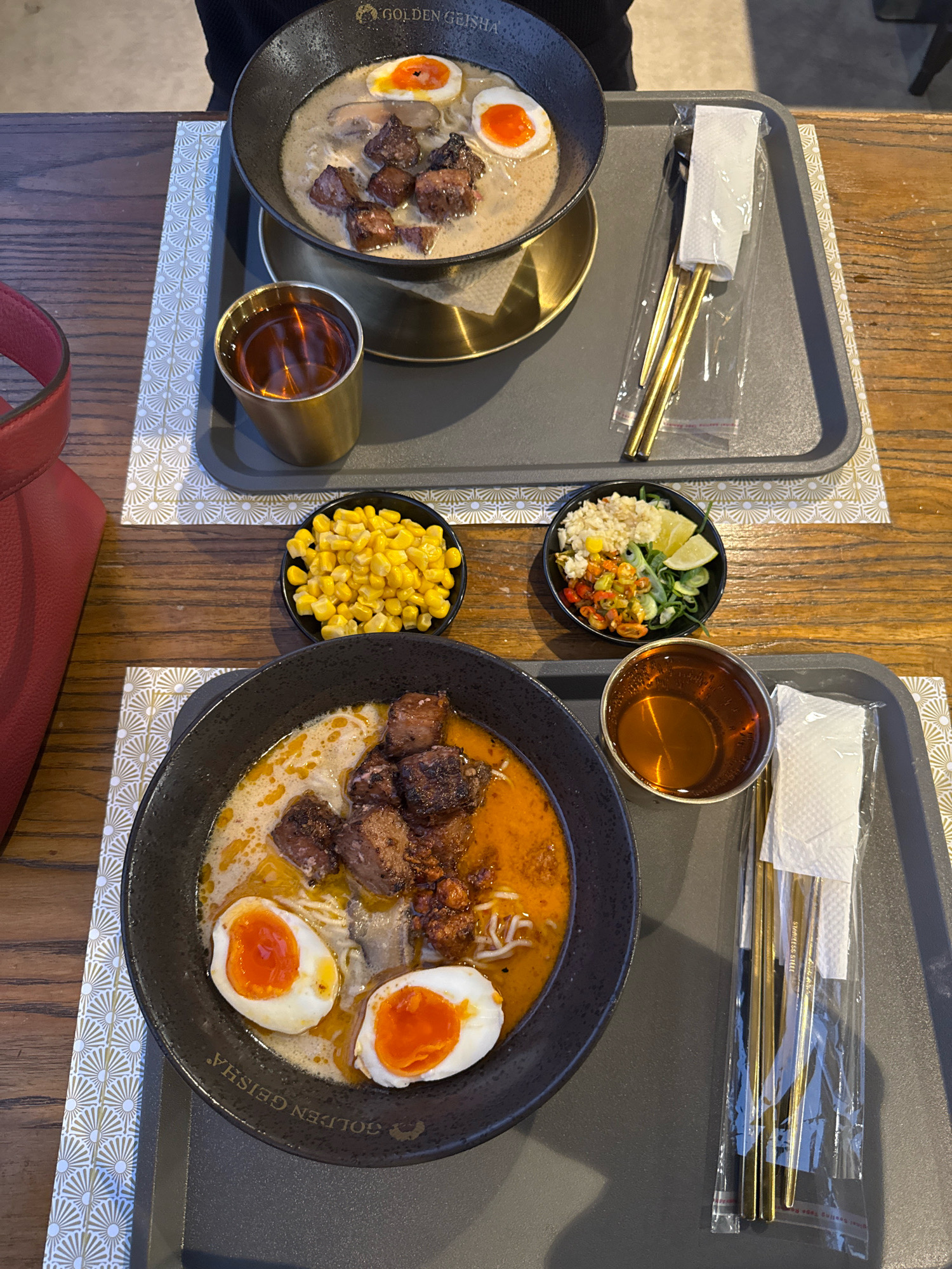 This is a top-down, medium shot image of two bowls of ramen on a wooden table, set on grey trays with golden chopsticks and napkins. The ramen bowls are black and appear to contain broth, noodles, meat chunks, and halved soft-boiled eggs. The top bowl has mushrooms as well. To the right of the top bowl, there is a golden cup with a dark liquid, likely tea. To the bottom left of the top bowl, there is a small black bowl of corn. To the bottom right of the top bowl, there is a small black bowl with chopped ingredients, possibly chili peppers, lime, and herbs. The bottom bowl has a similar presentation to the top bowl. There is a red bag on the left side of the image. The ramen bowls have "GOLDEN GEISHA" written on them. The image does not provide any information about the specific location within the city of Semarang, Indonesia.