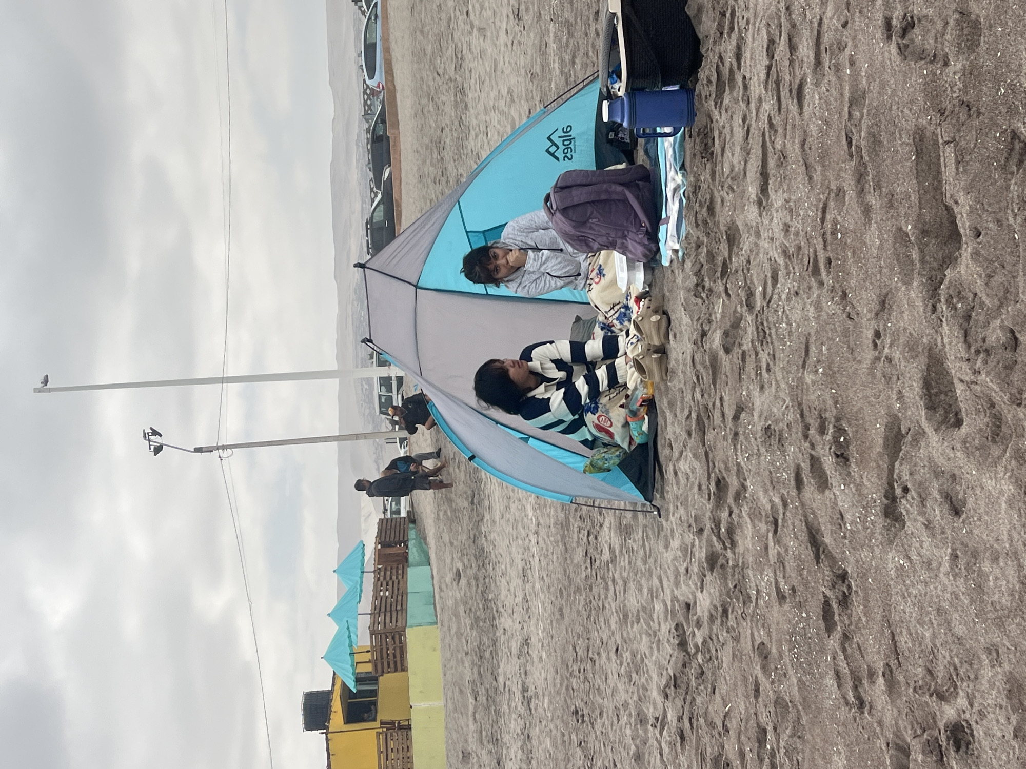 The image shows a beach scene in Arica, Chile, characterized by dark sand. In the foreground, there is a small, pop-up tent with two people inside. One person is wearing a striped shirt, and the other is in a light-colored sweatshirt. Beside the tent are various belongings, including bags and water bottles. In the background, rows of colorful stacked wooden structures are visible, interspersed with blue parasols. Further back, people walk along the beach near parked cars. The sky overhead is overcast.