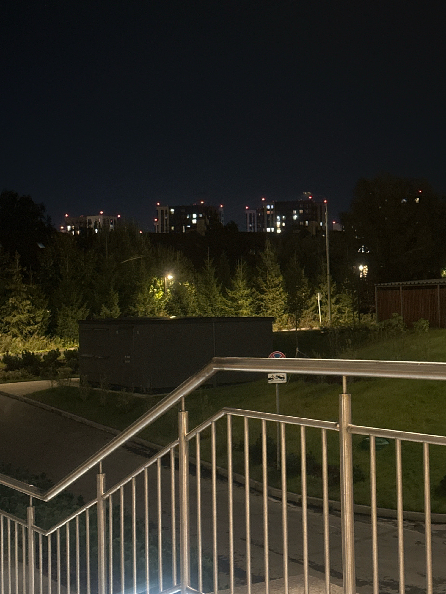 The image depicts an exterior scene at night in the city of Kommunarka, Russia. In the foreground, a metal handrail and vertical bars of a staircase are visible, reflecting ambient light. Beyond the staircase, a paved pathway leads towards a grassy area with some scattered vegetation. In the mid-ground, a dark, rectangular structure, possibly a utility building or storage unit, is situated amidst trees. A streetlamp with a bright yellow light illuminates the surrounding foliage. Further in the background, several multi-story buildings with illuminated windows and red lights on their rooftops are silhouetted against the dark night sky. The overall environment appears to be a residential or urban area with park-like elements.