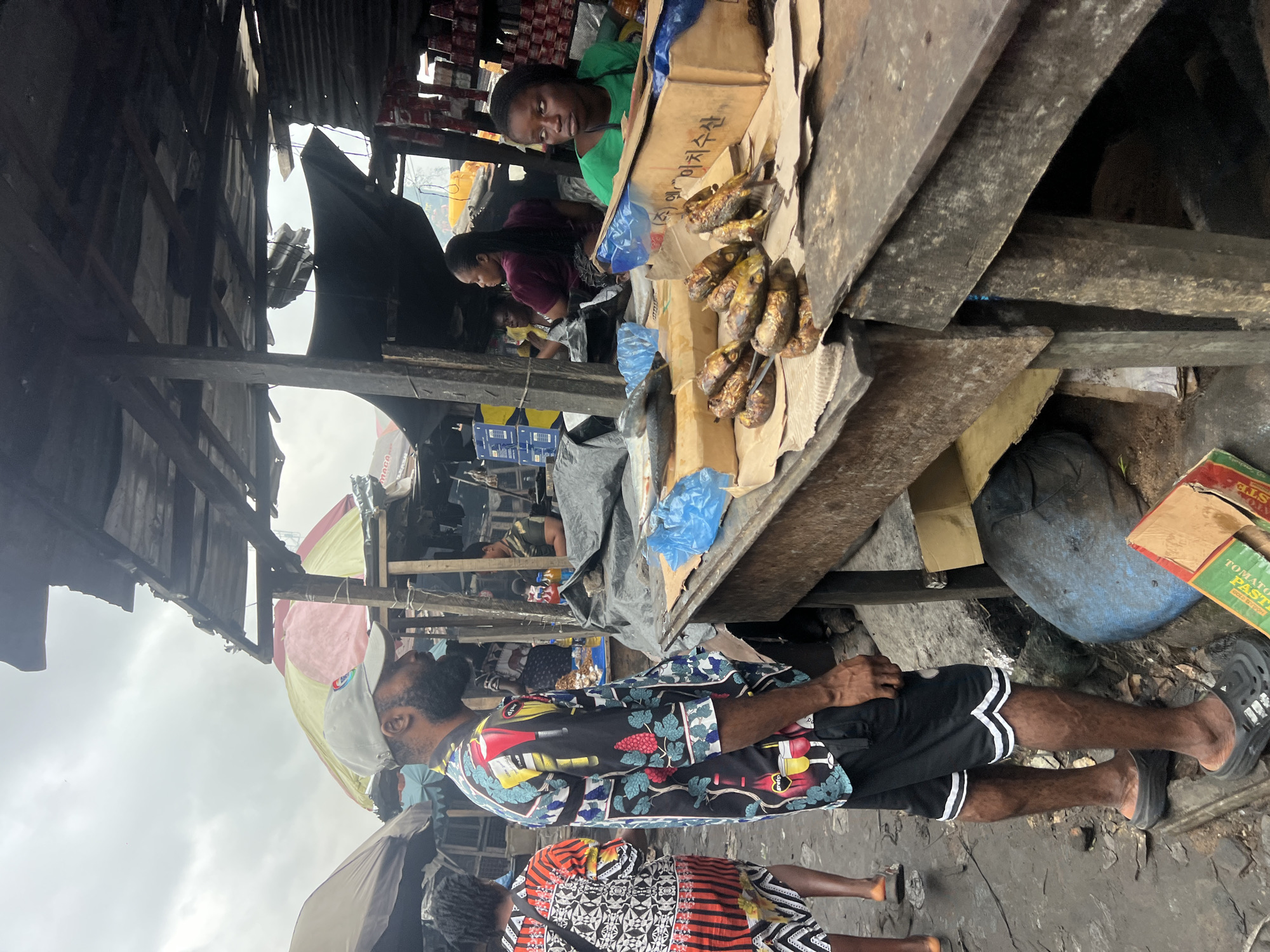 The image depicts a bustling outdoor market scene in Nkpor, Nigeria. In the foreground, a man with a beard wearing a grey baseball cap and a vibrant, patterned shirt stands facing to the right, his left side towards the camera. He is wearing black shorts with white trim and dark grey clogs. To his left, a woman with braided hair, dressed in a colorful, patterned dress, walks away from the camera.
The market stalls are constructed from rough wood and corrugated metal, with tarpaulin coverings providing shade. A wooden table in the midground displays various items, including several dried fish laid out on paper, some wrapped in blue plastic, and two larger fish in the background. A cardboard box with Korean writing is visible on the table, with more fish displayed. Behind the table, a young woman wearing a green top is seated, looking to her right. Further back, other individuals can be seen amidst the market stalls. The ground is uneven and appears to be dirt and debris, indicative of an active and somewhat unkempt market environment. The sky is overcast, suggesting a cloudy day.