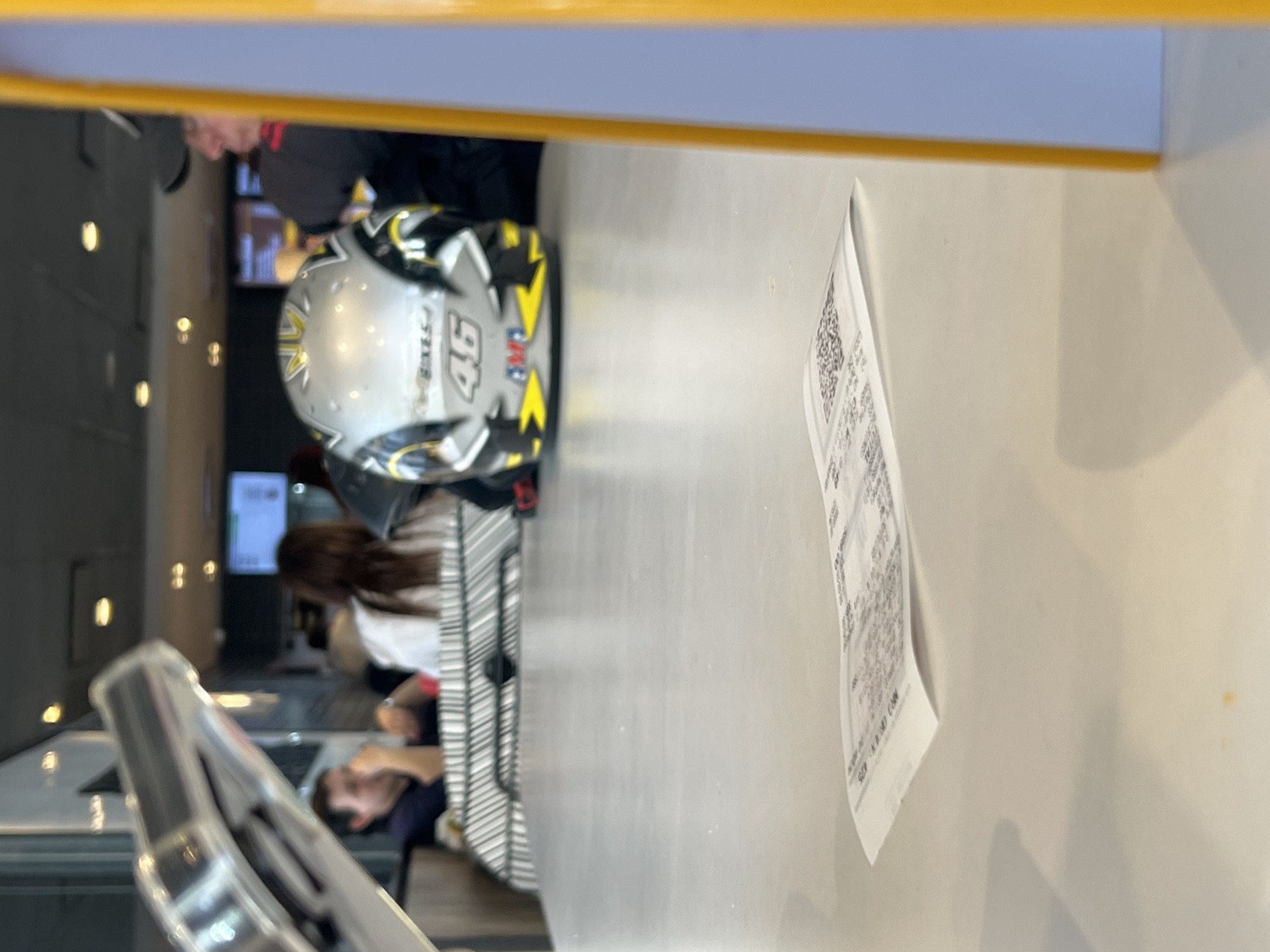 This indoor photograph, if rotated correctly, captures a casual setting, likely a cafe, restaurant, or service area, with a counter or table dominating the foreground. A prominent object on this light-colored surface is a distinctive motorcycle helmet. It is silver or light gray with bold black and yellow lightning bolt graphics. Clearly visible text on the helmet includes "VR46 BIKES" and the large number "46," strongly associating it with famous motorcycle racer Valentino Rossi.
To the right of the helmet, a white paper, appearing to be a receipt, stands upright. It displays tightly packed, mostly illegible print and a QR code. Closer inspection reveals fragments of text such as "NR3 ORD CAN" and the number "475."
In the blurred background, several people are visible. Towards the upper left, a person wearing a dark cap and dark clothing is partially seen. Further back and to the right, a person with long dark hair, dressed in a white top, is seated, possibly on a bench. In the lower left part of the background, a man with short dark hair is also seated, appearing to be looking down at something on a table. The background features various blurred lights, indicating an active, well-lit indoor environment. The scene suggests people are waiting, dining, or socializing in a place with a connection to motorcycle culture.