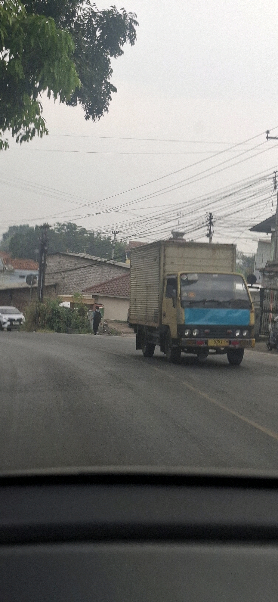 This is a medium-distance, eye-level, vertical shot of a yellow truck with a light blue front panel driving on a paved road in Sirnagalih, Indonesia. The truck is on the right side of the road, moving away from the viewer. The truck has a corrugated metal cargo box. The road has a single yellow line marking the lane. To the left of the road, there is a white car, a person in dark clothing walking away from the viewer, and some greenery. In the background, there are buildings with red tile roofs, trees, and numerous overhead electrical wires. The sky is overcast and light gray.
