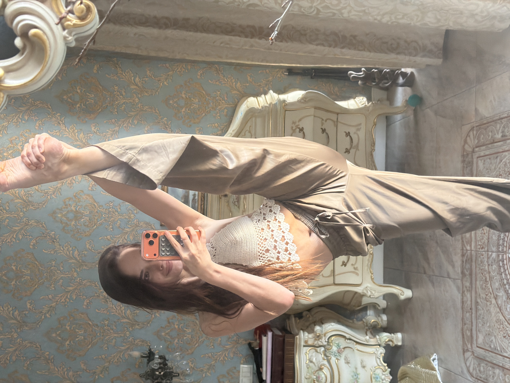 A young woman with long, dark hair is standing in a room, holding a phone to take a mirror selfie. She is wearing a white crocheted halter top and loose, tan pants. Her right leg is extended straight up in the air, her toes pointed. She is smiling and looking at the camera. The room appears to be a bedroom or dressing room, with ornate furniture and patterned wallpaper. The setting is consistent with a residential interior. There is no information in the image to suggest it is located in Ufa, Russia.