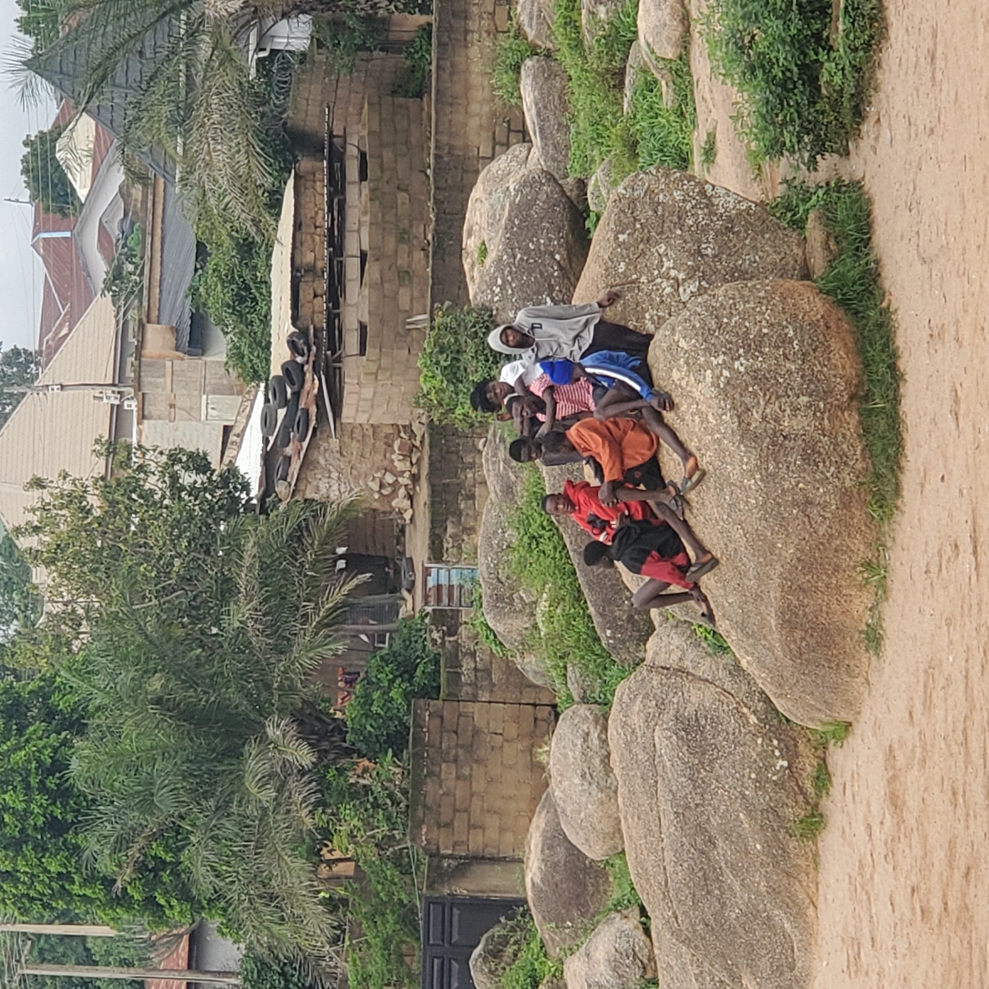 This image depicts a group of nine young males gathered on and around large rocks, with a backdrop of brick buildings and vegetation. The scene is set in a dusty, outdoor environment characteristic of Jos, Nigeria. The individuals are dressed in casual attire, including hoodies, t-shirts, and pants, with some wearing sandals or flip-flops. They appear to be interacting with each other, with some seated on the rocks and others standing. The rocks themselves are varied in size and texture, with patches of grass growing between them. In the background, there are brick structures, some with corrugated metal roofs, and abundant green trees and bushes. The overall impression is a candid snapshot of a group of youths in a natural, rocky area within a populated urban or semi-urban setting.