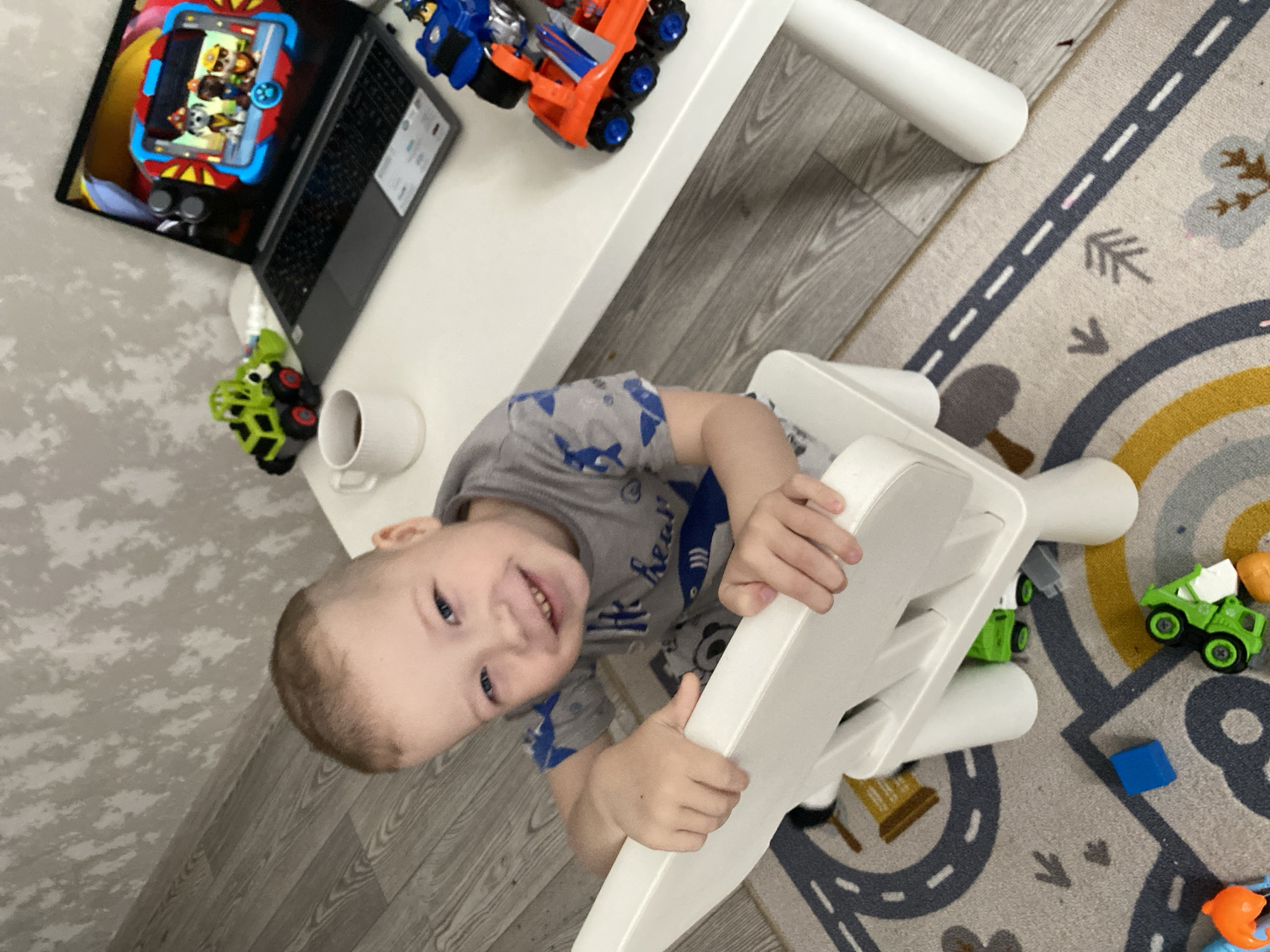A Caucasian boy with short brown hair and blue eyes sits on a white chair in what appears to be a playroom or child's room. He is wearing a gray t-shirt with blue shark designs. He smiles at the camera while holding the back of the chair.
On a white table to his left is an open laptop displaying a cartoon image on the screen, alongside several toy trucks in blue and orange. A white mug and a small green toy truck are also on the table.
The floor is covered with a carpet featuring patterns of roads, clouds, and rainbow arcs. Other toys, including more small green trucks and a blue block, are scattered around the base of the chair.
The location is unknown and there is no verifiable information that the image was taken in Sochi, Russia.