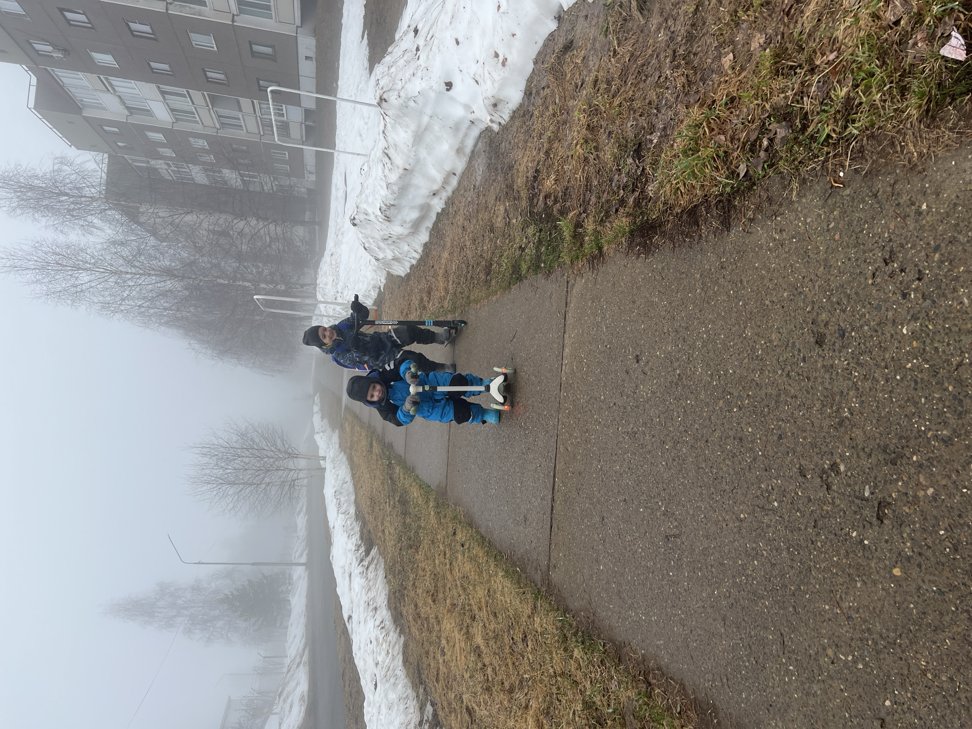 Two children are riding scooters on a sidewalk in a residential area in Markovskiy, Russia. It is a foggy day. The building is in the background, along with trees and snow. The edge of the sidewalk has grass and dead vegetation.