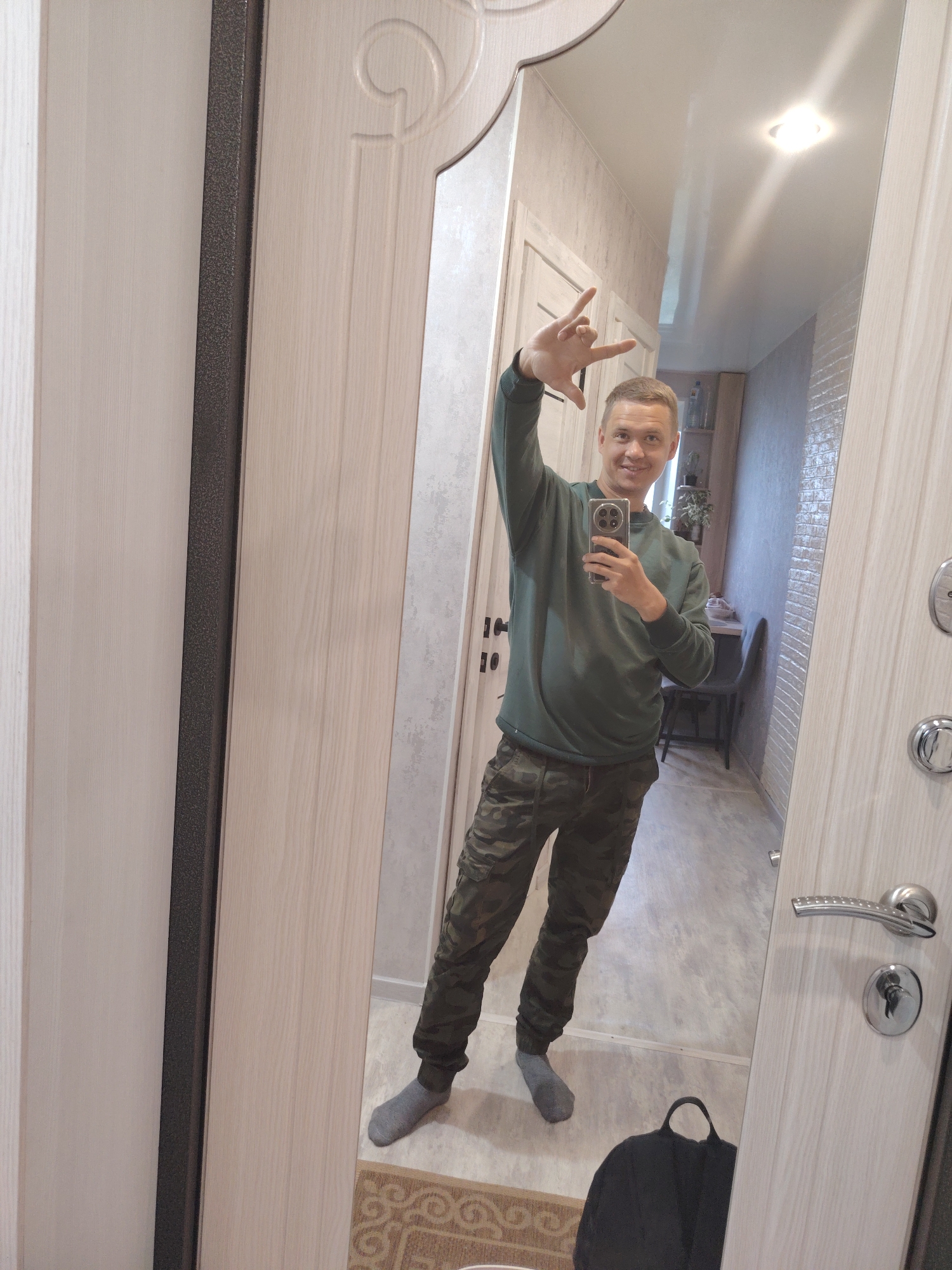 A man is taking a mirror selfie in what appears to be a hallway inside a residence in Chaikovsky, Russia. The full-length mirror is integrated into or mounted on a light-colored wooden door with decorative carvings at the top. The man, a Caucasian individual with short, light-colored hair, is smiling warmly at the camera. He is wearing a dark green long-sleeved top, camouflage-patterned trousers, and grey socks. His right hand is raised, making a gesture with his thumb, index, and pinky fingers extended, commonly known as a "rock and roll" or "I love you" sign. He is holding a smartphone with a visible multi-lens camera array in his left hand, using it to capture the image.
The reflection in the mirror reveals a hallway behind the man with light-colored, possibly textured wallpapered walls and light wood-effect flooring. A ceiling light fixture illuminates the space. In the background reflection, there's a small table or shelf with a plant and other items, as well as a dark chair. Part of a patterned rug is visible on the floor near his feet. A black backpack is also visible on the floor, reflected in the lower left portion of the mirror. The door the mirror is on has a silver handle and lock, with keys inserted. The overall lighting suggests an indoor setting, possibly during the day. No visible text is present in the image.
