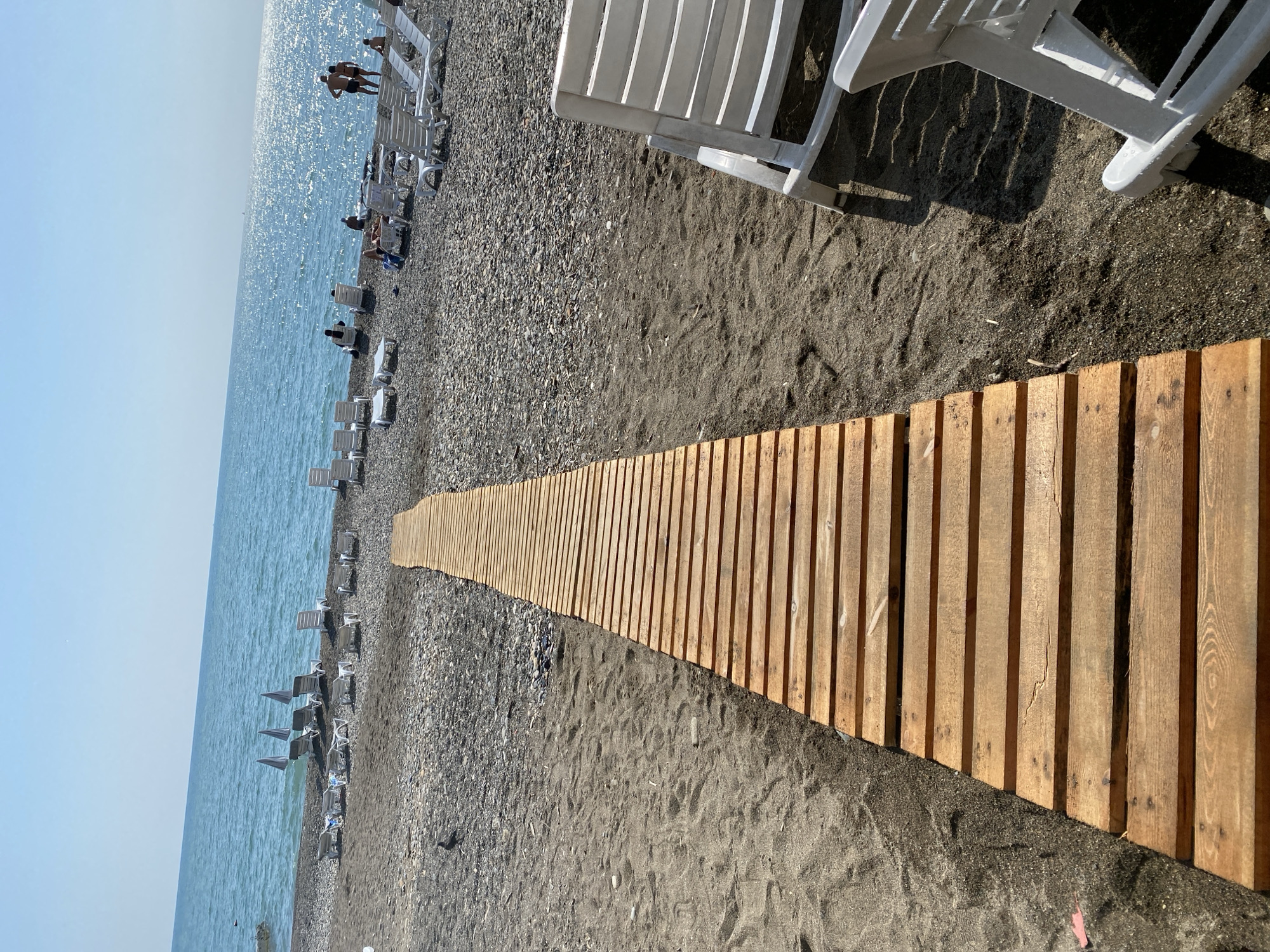 The image depicts a beach in Chabkwara, Abkhazia, on a sunny day. A wooden boardwalk extends from the foreground towards the sea, which is visible in the background. The beach is covered in dark sand and pebbles. Numerous white lounge chairs are scattered across the beach, some occupied by people. The sea is calm and sparkling under the bright sun. The sky is clear and blue. The overall scene is one of a typical summer day at a seaside resort.