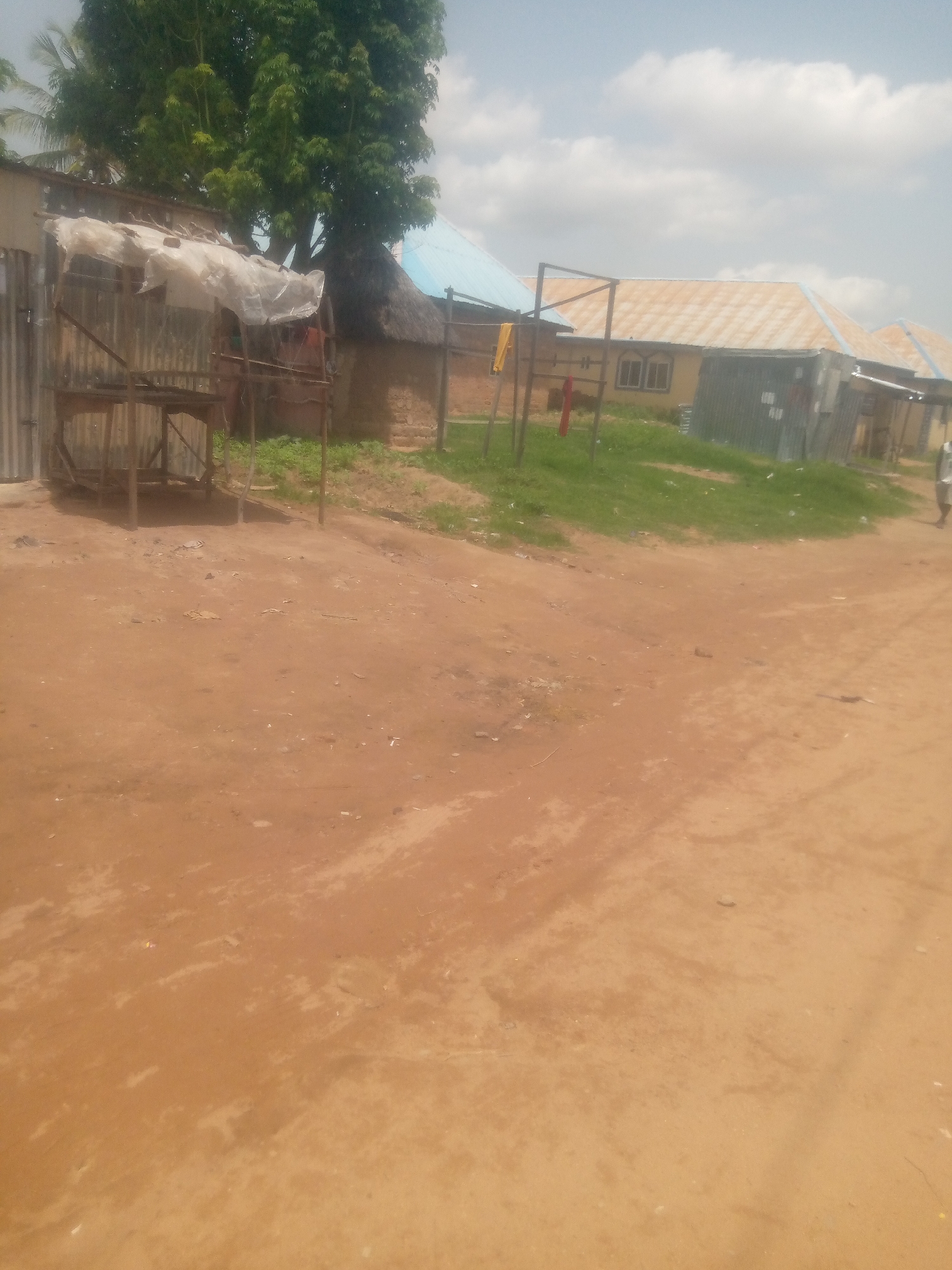 The image depicts a dusty, unpaved road in Jauro Yinu, Nigeria, with buildings and vegetation visible on the side. On the left, a makeshift stall constructed with corrugated metal and wooden planks is covered with a translucent sheet. Behind it, a large tree with dense foliage stands tall, partially obscuring a thatched-roof structure. Further to the right, a row of buildings with light-colored corrugated metal roofs and yellow walls line the road. One building has arched windows with grills. In the background, the sky is partly cloudy. A person is walking away from the camera on the right side of the road. The ground is predominantly red earth, with some patches of dry grass and scattered debris.