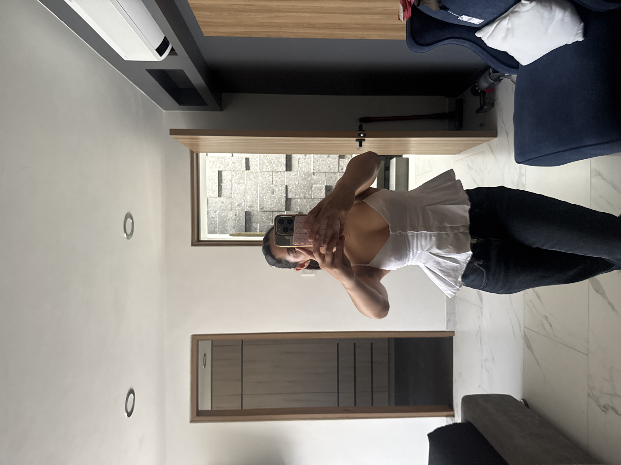 A young woman with shoulder-length black hair is taking a selfie in what appears to be a modern residential space. She is wearing a white peplum top and dark-wash jeans. She holds a smartphone with a light pink case in front of her face, obscuring it.
The environment includes a living room or bedroom area, judging by the presence of a dark blue armchair and a dark gray upholstered sofa, both placed on a white marble floor. The walls are painted in light shades. There are modern-style circular wall accents. A wooden-framed door leads to an outdoor space or another room, indicated by the view of what seems to be a textured wall. A wide door frame suggests an entrance to a hallway, with a closet or storage units behind it. An air conditioning unit is mounted near the ceiling.
Based on the architectural style, material finishes, and location information, the photo likely comes from a modern apartment or house within a newer development in Puebla, Mexico.