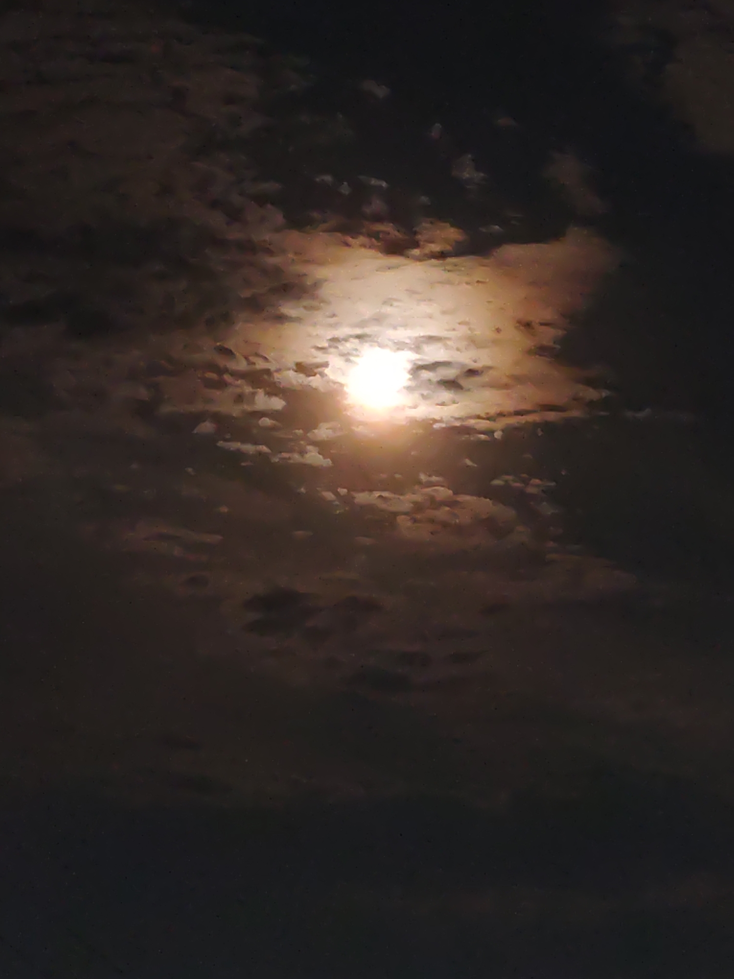 This is a nighttime photograph of a cloudy sky, with the moon visible through a break in the clouds. The moon is bright and illuminated, casting a warm glow on the surrounding clouds. The clouds are dark and textured, with some appearing lighter and more wispy where they are illuminated by the moon. The overall scene is dark and atmospheric, with a hint of color from the moon's light. There are no people, subjects, objects, or actions visible in the image. The location in the city of Petropavl, Kazakhstan is not discernible from the image itself.