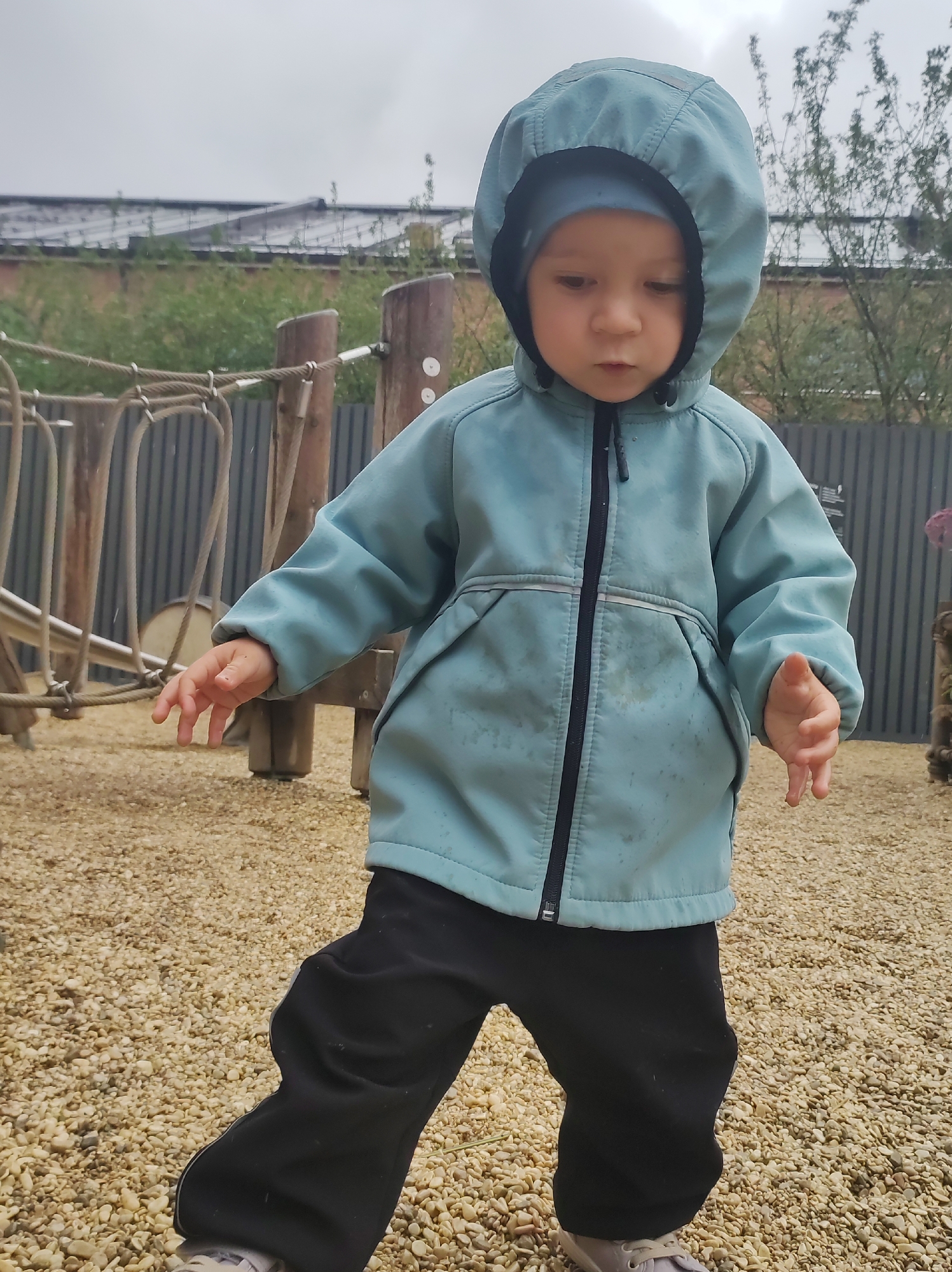 A young child, possibly a toddler, stands on a surface covered in light-colored gravel. The child is wearing a teal hooded jacket and black pants. The jacket is zipped up, and the hood is pulled over the child's head, obscuring part of their face. The child's hands are visible, with the left hand extended outwards. In the background, there is a fence, wooden structures, and some greenery. The sky appears overcast. The scene suggests an outdoor play area, possibly a playground. There is no visible indication of the child's specific location within Saint Petersburg, Russia.