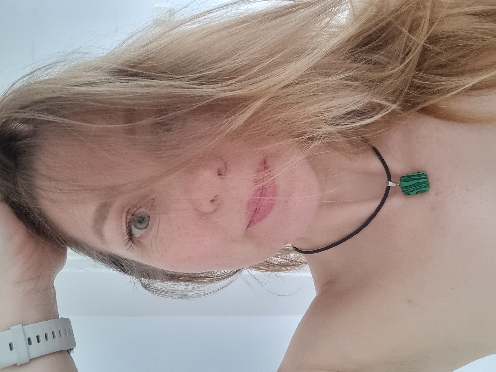 A woman with fair skin and blonde hair is positioned looking down at the camera. She is wearing a necklace with a green square pendant. Her left arm is raised, and she is wearing a white watch. The background appears to be a plain white surface. The image does not contain sufficient information to determine the location or any actions/interactions taking place.