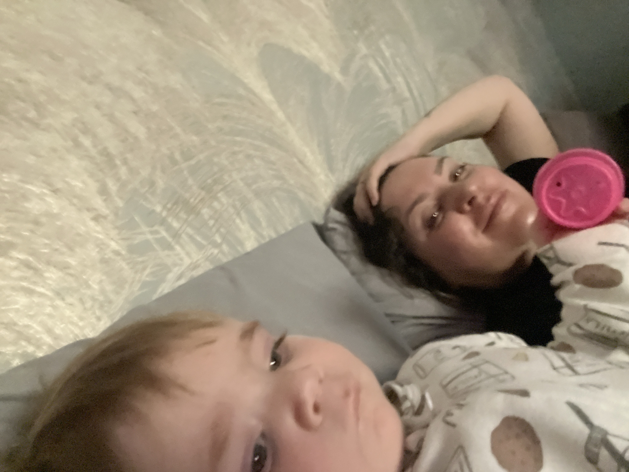 A selfie taken indoors features an adult and a child lying on a bed. The adult, positioned on the right side of the frame, is lying on a gray pillow with their head resting on a hand. They are holding a pink sippy cup with a star-shaped design on the lid. The child is positioned on the bottom left of the frame, lying on a gray pillow. Both are covered with a blanket patterned with images of pastries. The background includes a textured headboard or wall. The location is unknown.