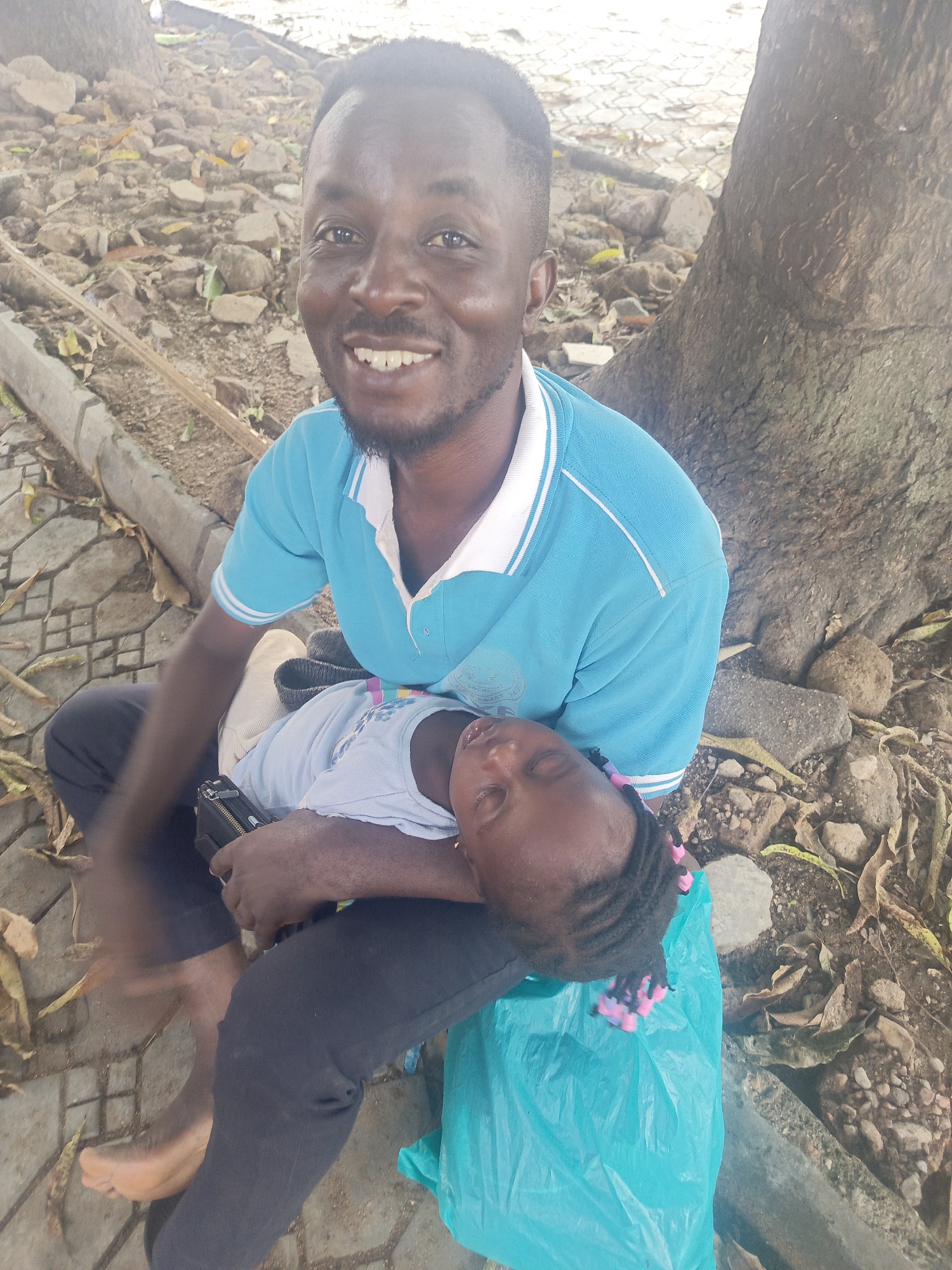 A man sits cross-legged on a paved area in Akwanga, Nigeria, with a sleeping child resting on his lap. The man wears a light blue polo shirt with a white collar, and dark pants. He is smiling and looking at the camera. The child is wearing a light-colored shirt, and has braids with pink decorations. The man holds a dark object, possibly a wallet or phone, in his hand. A turquoise plastic bag is on the ground next to the man. Behind them, the base of a large tree is visible, with fallen leaves and small stones scattered around it. A concrete curb borders the paved area.