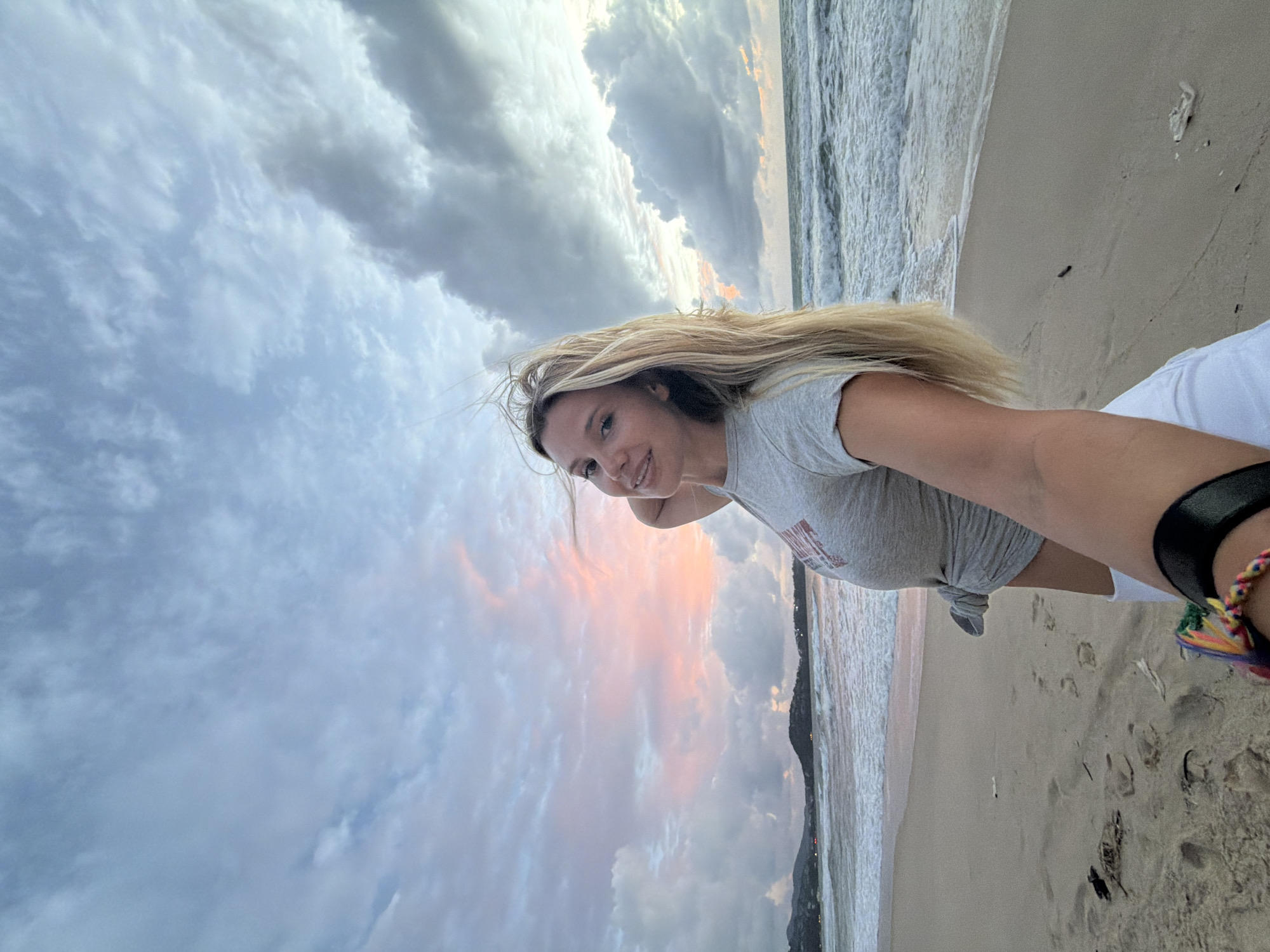 A blonde woman stands on a sandy beach in Choeng Thale, Thailand, during what appears to be sunset or sunrise. She is wearing a gray t-shirt tied at the waist and white shorts. The sky above is filled with clouds, some of which are tinged with pink and orange hues. The ocean is to her right, with waves breaking on the shore. In the background, hills and lights from a settlement are visible. The woman is holding the camera, taking a selfie, and her left arm is bent with her hand touching her head, her hair is blowing in the wind.