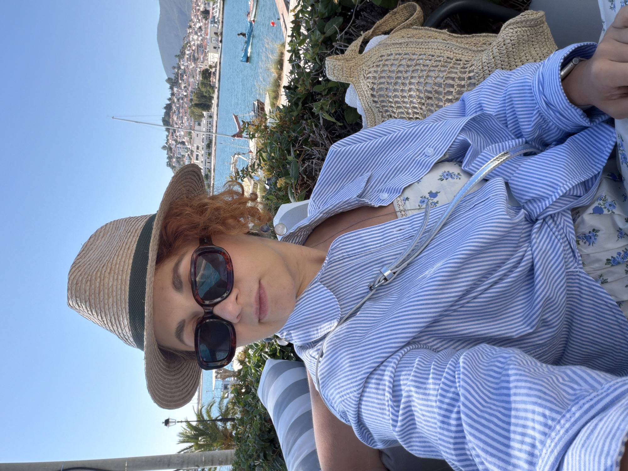 A woman is captured in a bright, outdoor, self-portrait-style shot, set against a picturesque coastal town. She is wearing a light straw hat with a dark band and large, dark-rimmed sunglasses. Her reddish, curly hair frames her face. She is dressed in a blue and white striped, long-sleeved button-down shirt, worn open over a white top with a delicate blue floral pattern. A slender necklace and a light-colored strap (possibly from sunglasses or a bag) are visible across her chest. She appears to be seated comfortably, with a striped cushion visible behind her.
To her right, a woven straw bag rests. The background reveals a stunning view under a clear, azure sky. A calm body of water, likely a sea or bay, features a few boats, including the prominent mast of a sailboat. Beyond the water, a charming town with numerous white and light-colored buildings stretches up a hillside, backed by majestic mountains. Green foliage, including bushes and palm trees, adds to the vibrant outdoor setting. The bright sunlight indicates it is daytime, suggesting a relaxed moment in a scenic location like Poros, Greece. No text is visible in the image.