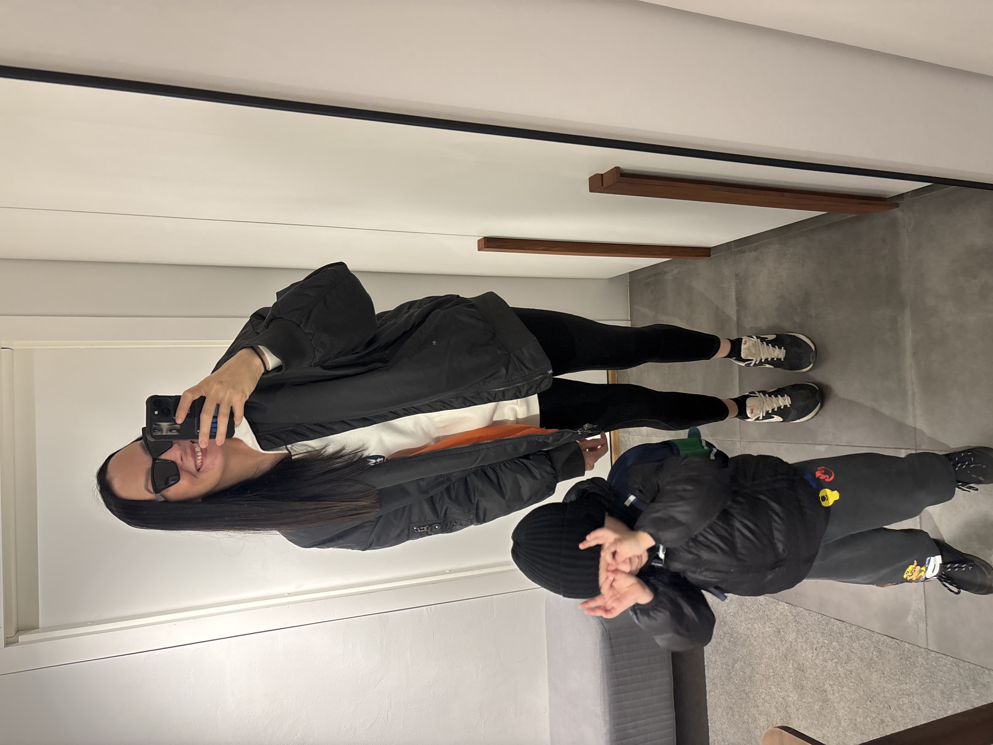 A woman with long dark hair and sunglasses holds a phone up to take a mirror selfie with a young child. The woman is wearing a black jacket over a white shirt, black leggings, and athletic shoes. The child is wearing a black jacket, dark gray sweatpants with colorful cartoon patches, a black beanie, and dark-colored shoes. The background shows a dressing room with white walls, a gray floor, and wooden shelves. The location is unknown; there is no definitive evidence in the image that places it in Chaikovsky, Russia.