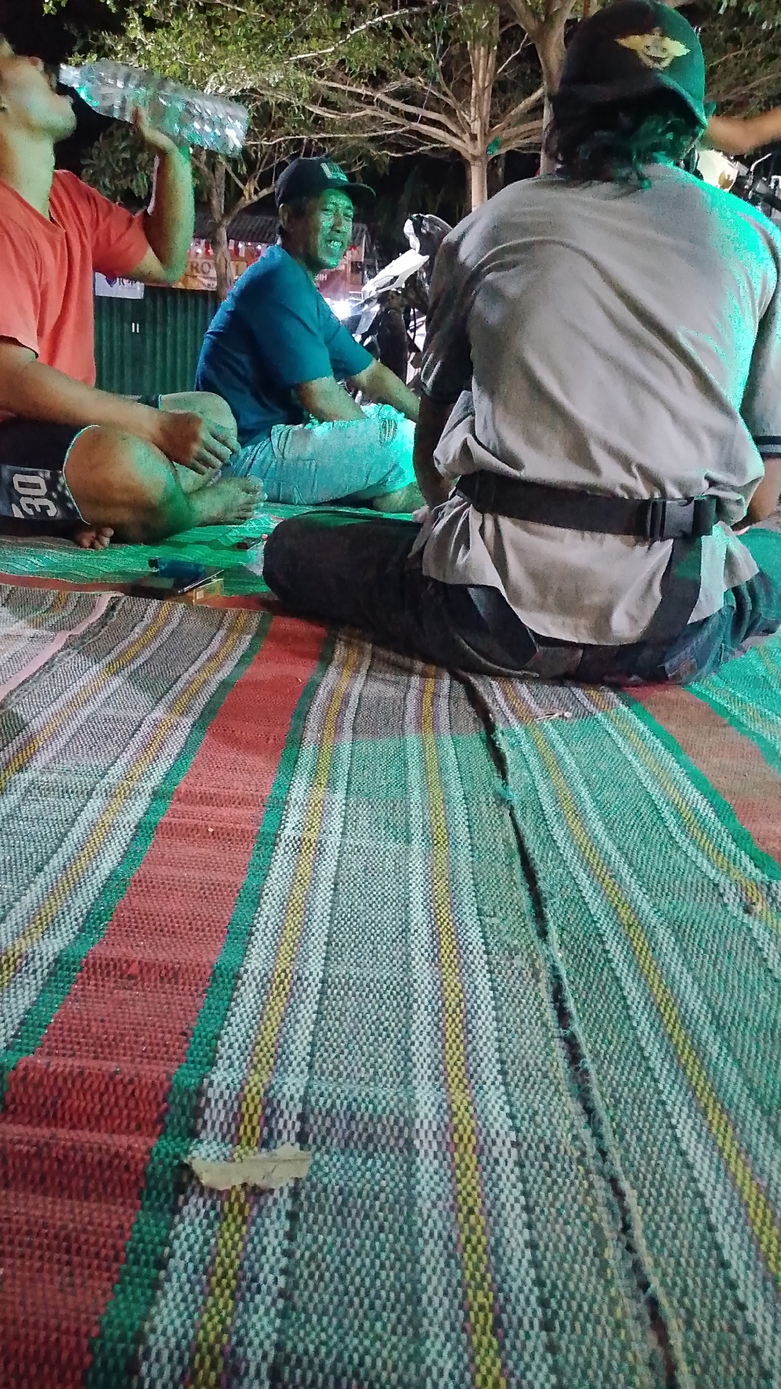 Three individuals are seated on a striped mat outdoors, likely at night in Karanglangu, Indonesia, illuminated by green artificial light. One person, wearing a red t-shirt, is drinking from a large plastic water bottle. To their left, another individual in a blue t-shirt and jeans smiles broadly. A third person, facing away from the camera and wearing a grey shirt with a black belt and a cap, is seated to the right. The background features trees and structures, hinting at an outdoor gathering or event.