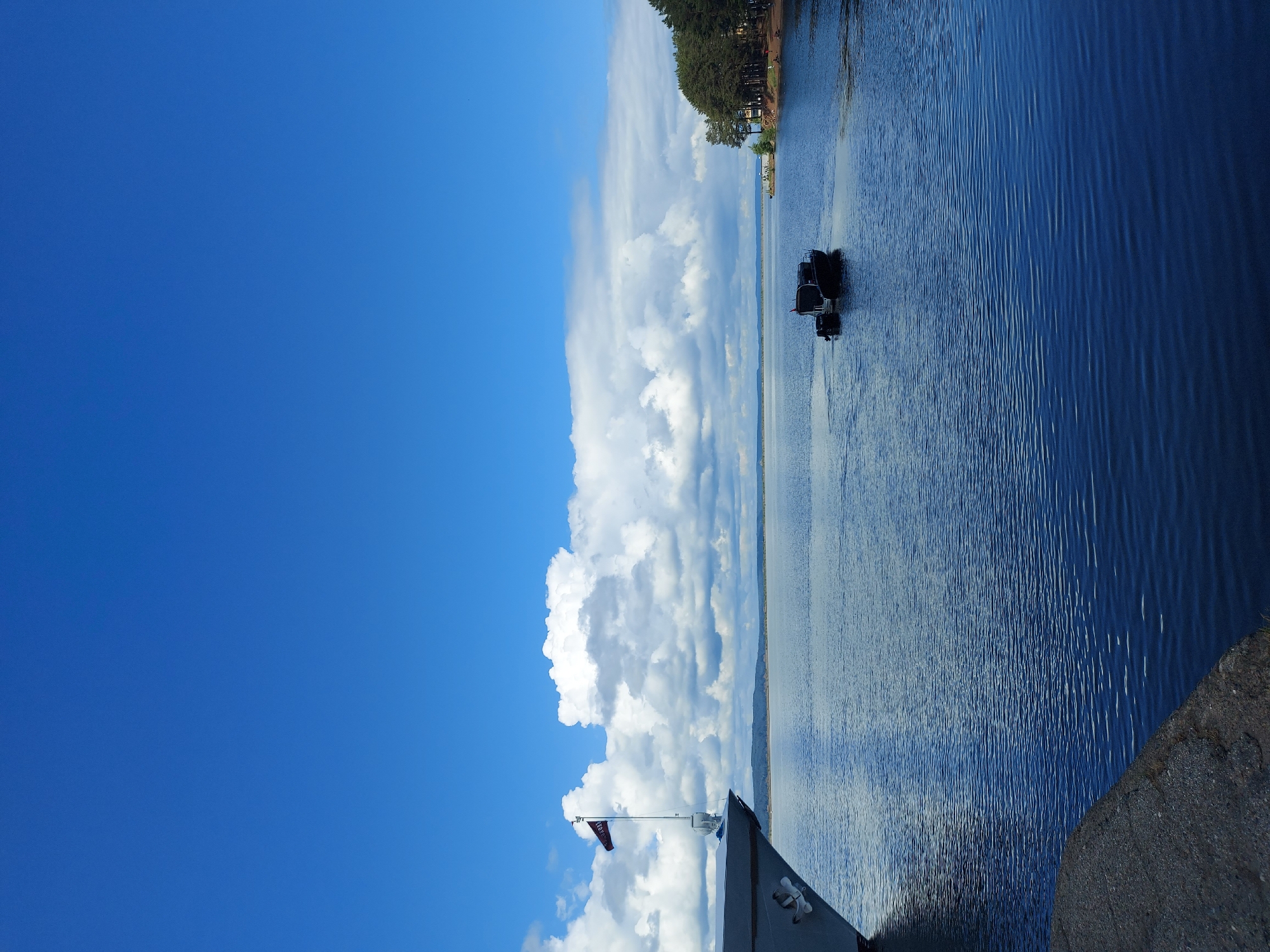 A sideways-oriented view over a large body of water, likely the Kama River, at Chaikovsky, Russia. The photograph is taken from an elevated position, possibly the deck of a structure or boat. The sky dominates the left side of the frame, showcasing a vibrant blue backdrop filled with billowing cumulus clouds. A flag mounted on a pole from the near foreground indicates the camera's location. The water extends into the background where its glassy surface reflects the sky, creating a sense of depth. A forested shoreline is visible along the distant edge of the body of water. There are two dark boats, connected to each other in the water. A small section of concrete structure is visible in the lower right-hand corner.