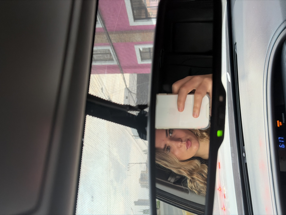 The image depicts a first-person view from inside a stationary vehicle, looking through the windshield and into the rearview mirror, which serves as the primary subject. The time displayed on the vehicle's digital dashboard clock is "6:17." An "AIR BAG OFF PASSENGER" indicator light is also visible on the dashboard. A green indicator light illuminates the bottom edge of the rearview mirror housing. Several small, red circular objects are present on the dashboard beneath the windshield.
In the rearview mirror, a female individual with long, wavy blonde hair is reflected. She is observed with a pouting expression, holding a white smartphone in her right hand, positioned to capture a selfie reflection in the mirror. The smartphone is enclosed in a clear or translucent case.
Through the vehicle's windshield, an urban environment in Puebla, Mexico, is visible. The sky is partially cloudy with a light hue, suggesting either early morning or late afternoon. To the left, distant multi-story buildings and utility poles are discernible, indicating a developed area. To the right, a prominent two-story building with a vibrant fuchsia-pink facade, contrasting white window frames, and a brown base section is clearly visible. Utility wires traverse the upper part of the scene against the sky. A fine mesh pattern, likely from a sunshade or windshield tinting, covers much of the upper external view.
