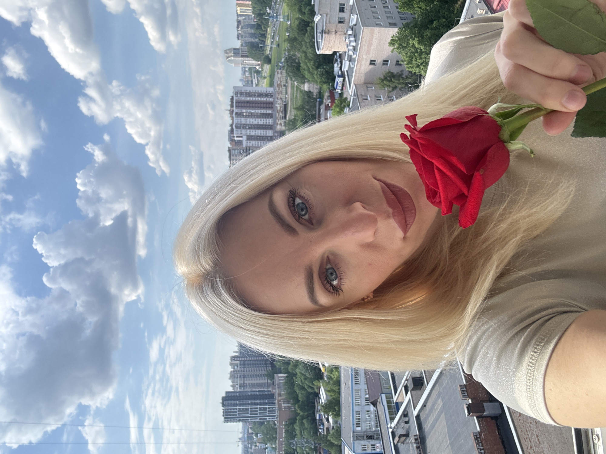This vertically oriented image is a portrait of a woman holding a red rose, set against an urban backdrop under a cloudy sky. The subject is a woman with long, blonde hair and light-colored eyes, looking directly at the camera with a calm expression. She is wearing a light-colored top and holding a single, vivid red rose with green leaves in her left hand, positioned near her face.
The background reveals a expansive cityscape, viewed from a high vantage point, suggesting the photo was taken from a building balcony or rooftop. Numerous multi-story buildings and high-rises are visible, interspersed with green trees and what appear to be parks or green spaces. Above the city, the sky is a bright blue with scattered white, cumulus clouds.
The image captures a sunny day, likely midday or afternoon, given the clear and bright lighting. The overall mood is serene. There is no visible text in the photograph.