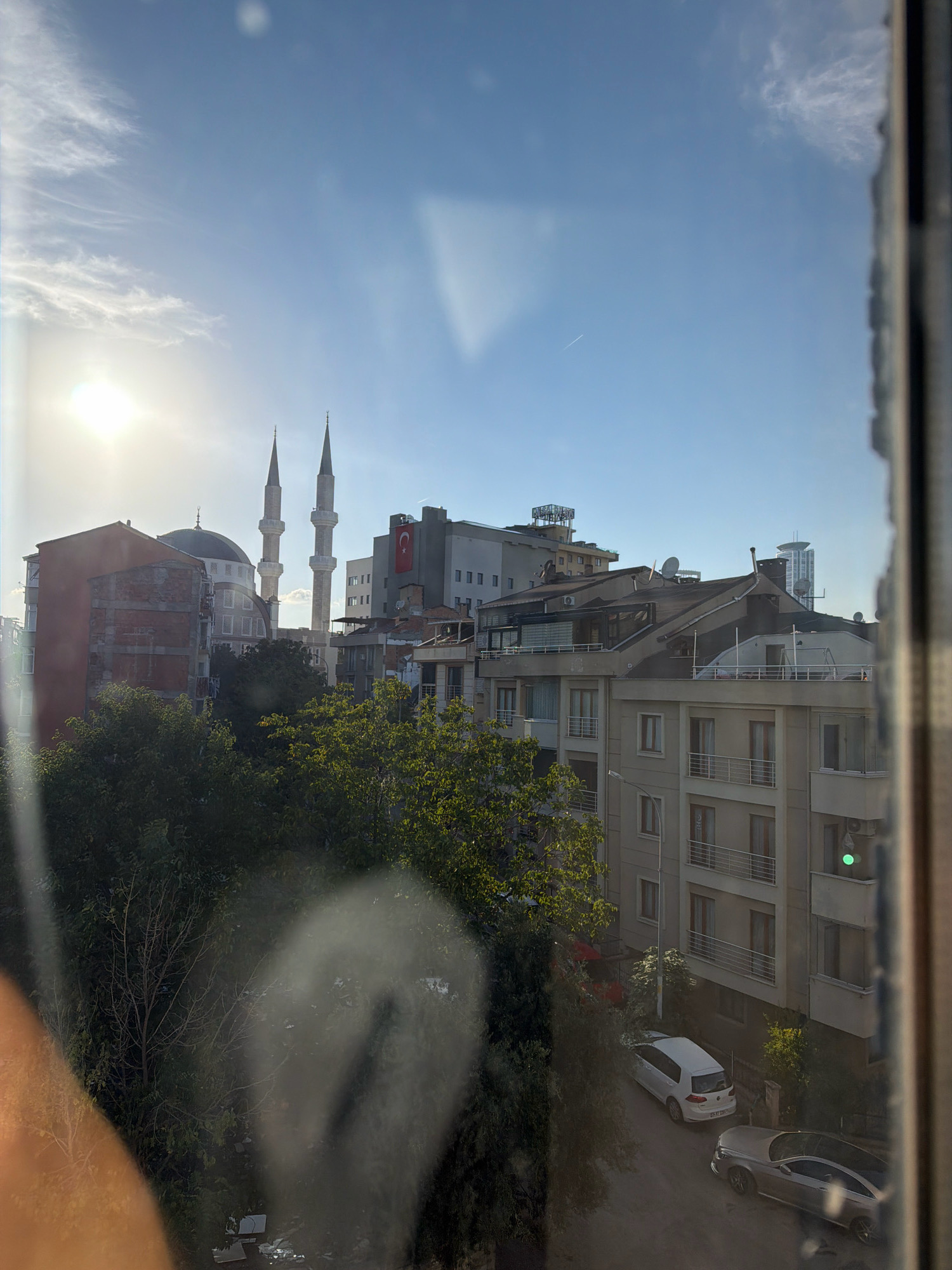 The image captures an urban scene in Ataşehir, Turkey, under a bright, partly cloudy sky. Dominating the cityscape are several buildings, including a mosque with two prominent minarets and a dome, its white structure contrasting with the surrounding architecture. Adjacent to the mosque is a building with a red-brick facade, appearing older and partially weathered. Other buildings in the mid-ground feature a mix of architectural styles, with balconies and rooftop satellite dishes visible. A Turkish flag is displayed on one of the taller buildings. In the foreground, dense green foliage from trees partially obscures the view of the buildings and a street below. Two cars are parked on the street: a white hatchback and a silver sedan. The sun is a bright, hazy orb in the upper left quadrant of the sky, casting a lens flare and illuminating the scene with a warm light. A faint contrail from an aircraft is visible high in the sky. Reflections on a windowpane are subtly present in the foreground, creating a layered effect in the image. The overall impression is that of a typical urban environment in Turkey, bathed in daylight.