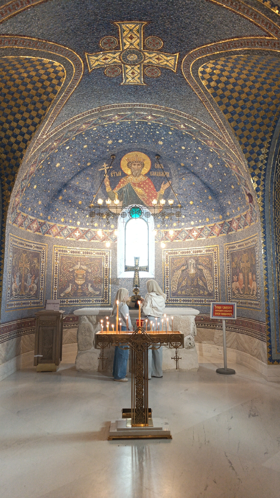 Two women are standing at a candle stand with lit candles, located in the interior of a church. The stand is a large cross-shaped structure made of gold metal. The church has mosaic walls and a mosaic ceiling with a large cross. A window can be seen in the background, along with a painting of a man holding a cross. A wooden cabinet is present on the left side. The location is the Church of St. Nicholas in Sevastopol, Ukraine.
