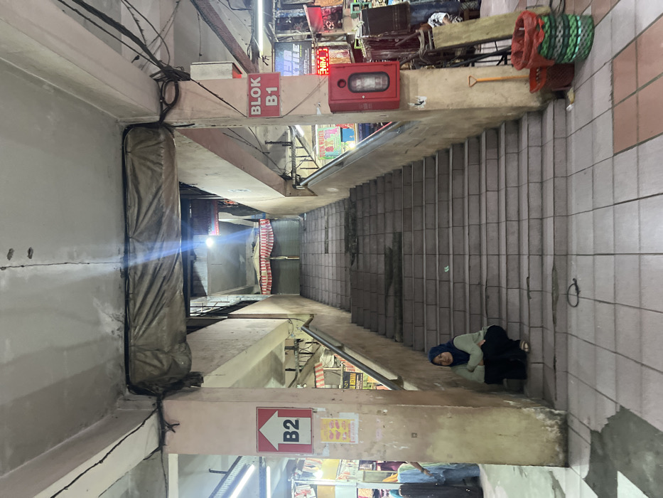 The image, oriented vertically on its side, provides a downward view into a multi-level market or shopping complex. A wide, dark grey tiled staircase descends prominently from the center towards the right side of the frame, leading to lower levels of the building. The architecture consists of sturdy concrete beams, pillars, and walls, with exposed electrical wiring and conduits running along the upper left.

Artificial lighting illuminates the scene, casting bright spots and revealing various shop signs and stalls in the background at different depths, indicating a busy commercial environment.

In the upper-middle section of the image, a red sign with white text "BLOK B1" and a right-pointing arrow is visible. Adjacent to it, a red fire extinguisher cabinet is mounted on a concrete beam, accompanied by a yellow-handled shovel and several red and green woven baskets. Further down, in the lower-middle, another red sign displays an upward-pointing arrow and "B2", suggesting different block or basement levels.

On the far right, near the bottom of the visible staircase, a person is seated on the tiled steps or floor. They are wearing a blue headscarf (hijab), a light-colored long-sleeved top, and dark pants, appearing to be resting or waiting. The setting has a utilitarian feel, with concrete and tiled surfaces, common in a bustling market environment in locations like Nganguk, Indonesia. The time of day is indeterminate due to the exclusive use of artificial interior lighting.